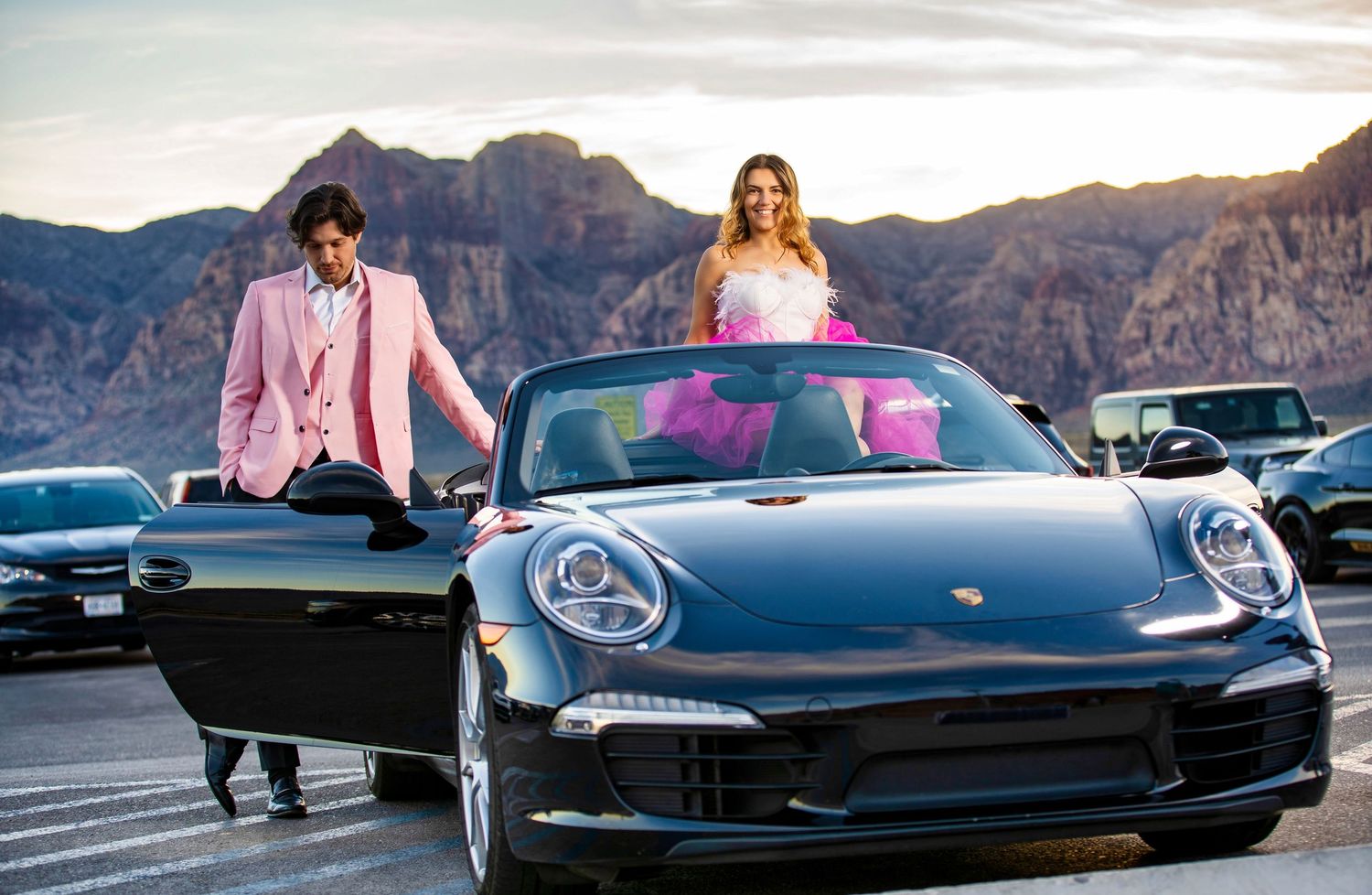 Couple in formal wear posing with a black Porsche 911 convertible at sunset with mountain backdrop.