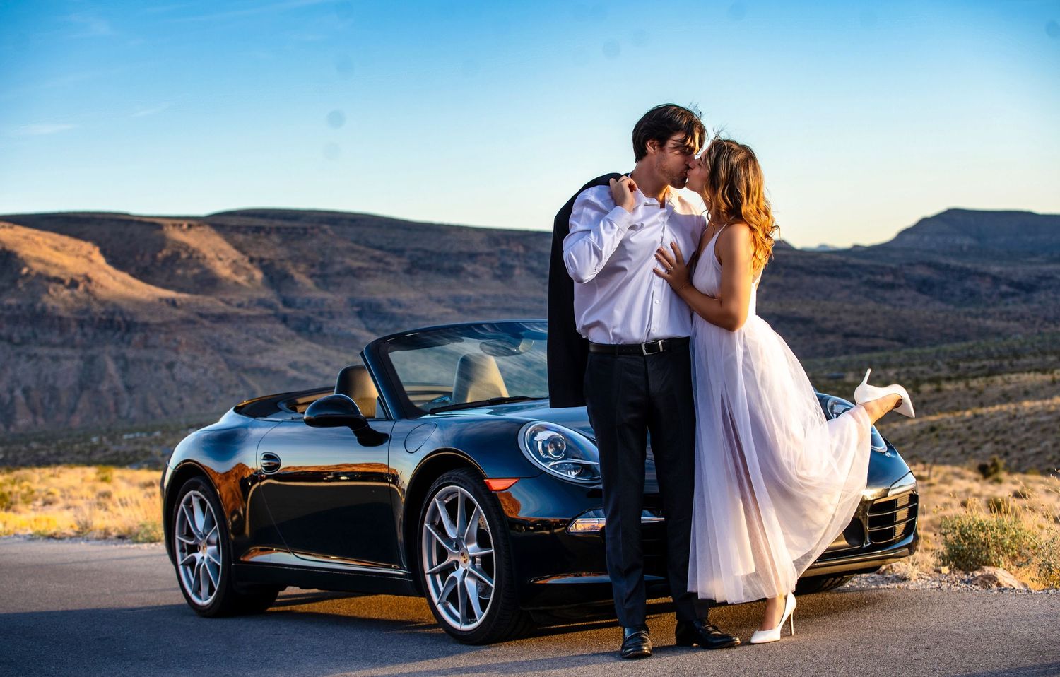 Bride and groom kissing beside a black Porsche 911 convertible with scenic desert mountains at sunset.