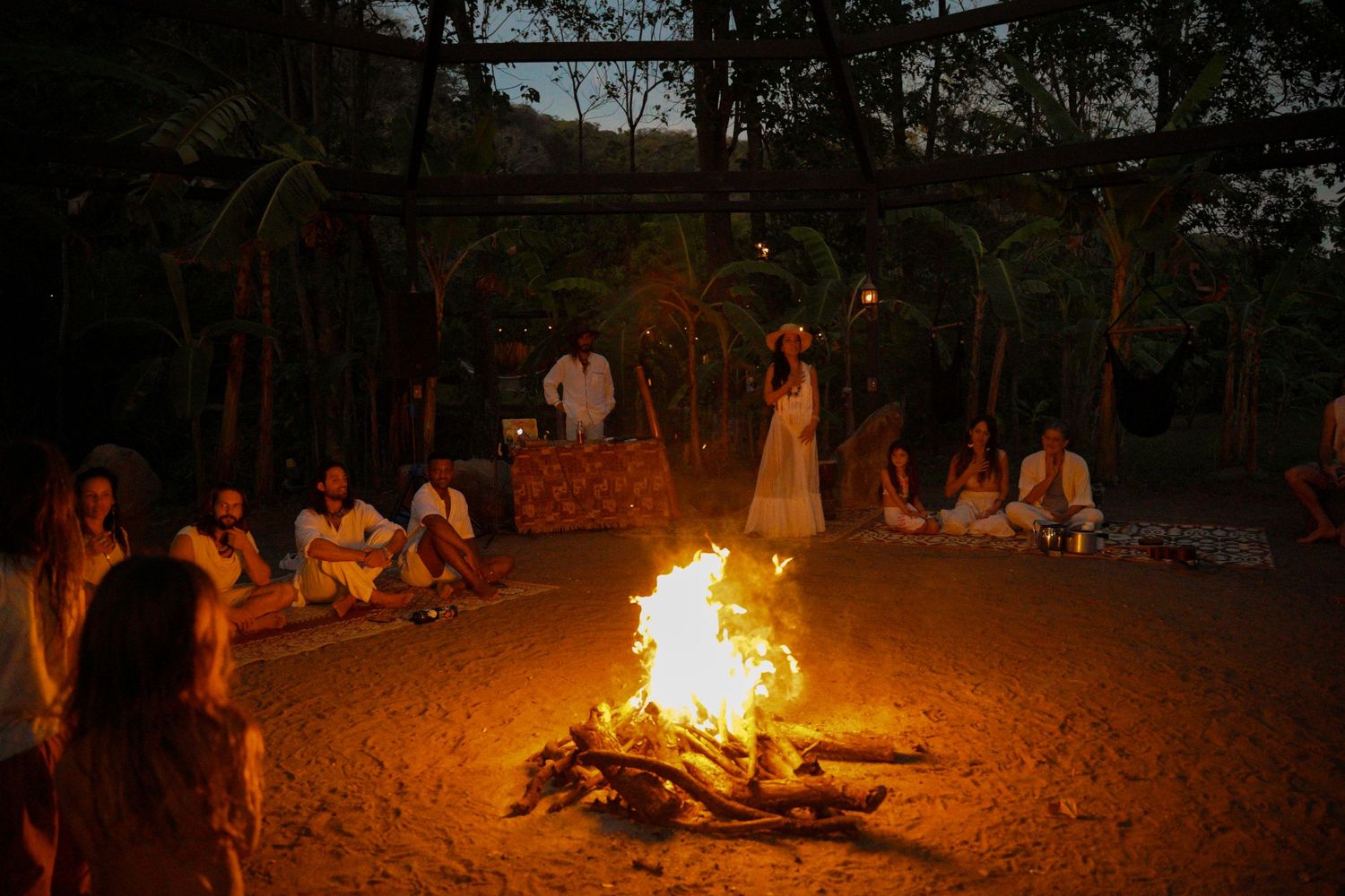 Group gathered around large bonfire at night in wooded outdoor setting.