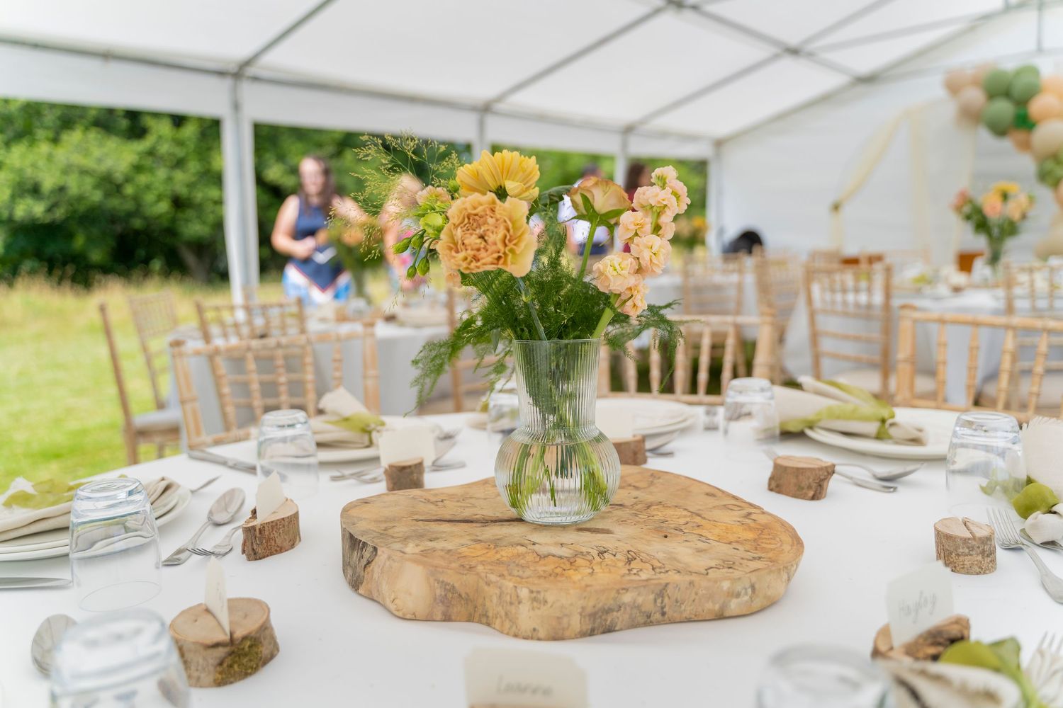 Elegant wedding table setting with rustic wood slice centerpiece holding yellow flowers in a glass vase under white tent.