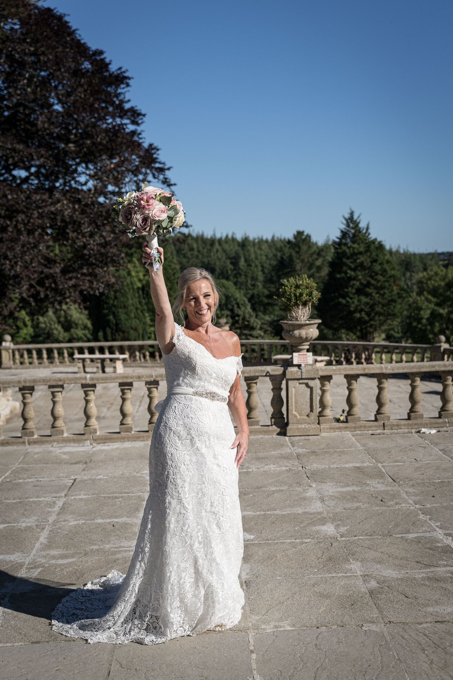 Bride in lace gown raises bouquet triumphantly on stone terrace with garden view. Elfordleigh Hotel Wedding. Devon Wedding Photography