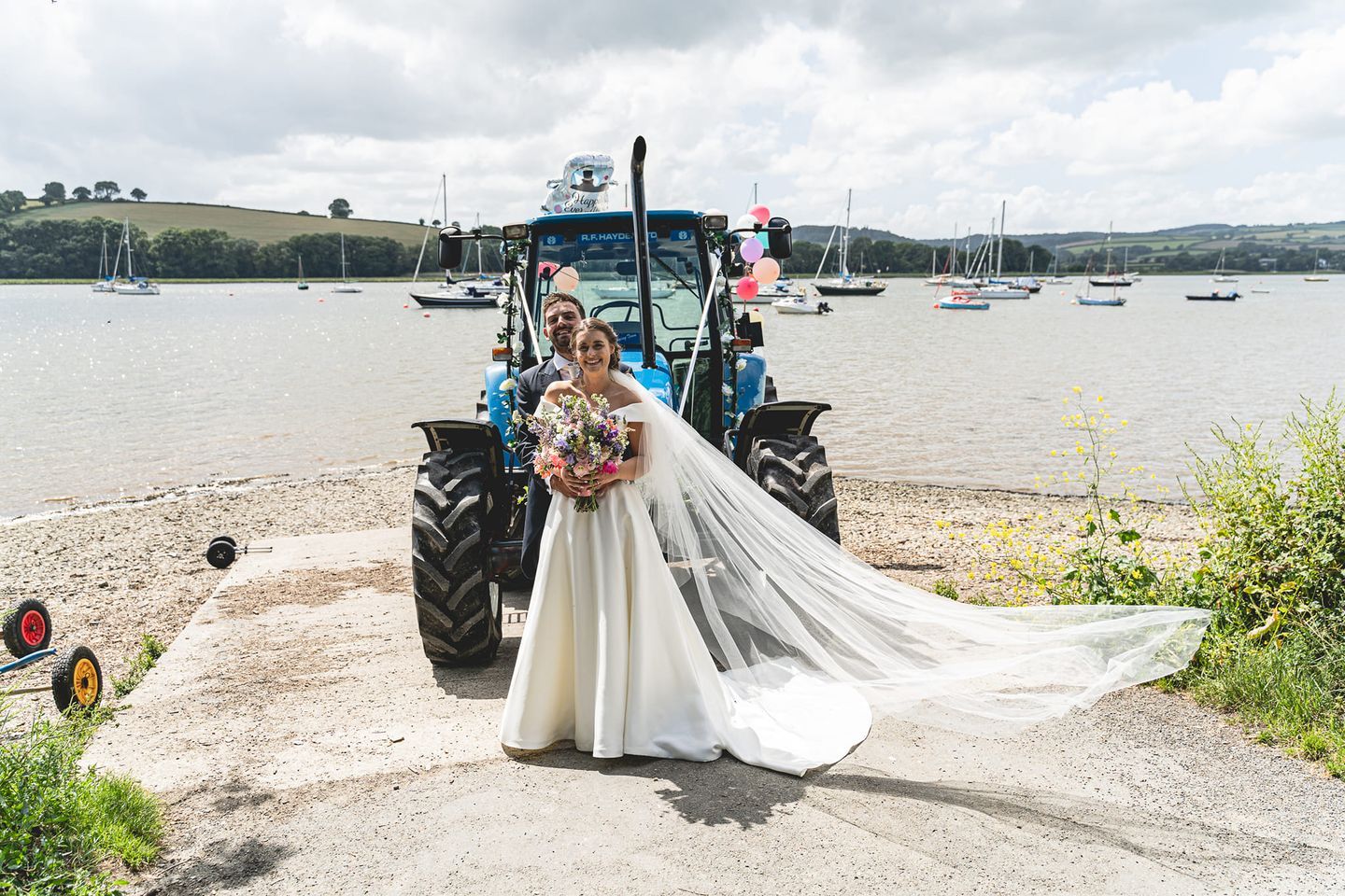 Bride in flowing white dress poses beside blue tractor at waterfront wedding location.