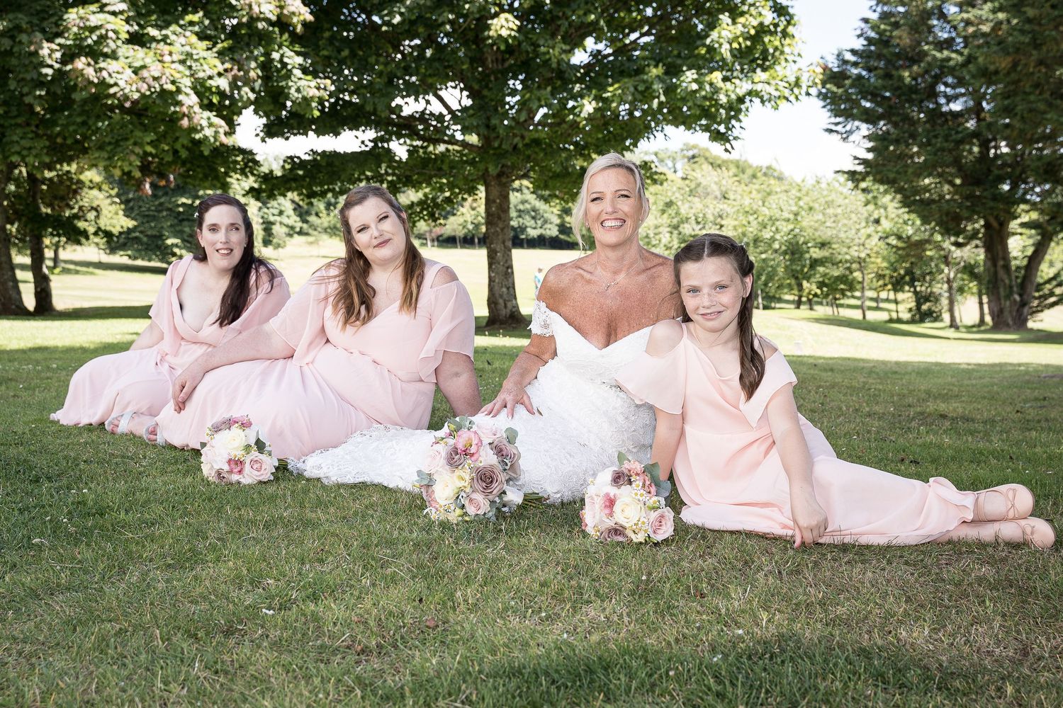 Wedding party in pink dresses relaxing on grass during outdoor photo session. Elfordleigh Hotel Wedding. Devon Wedding Photography