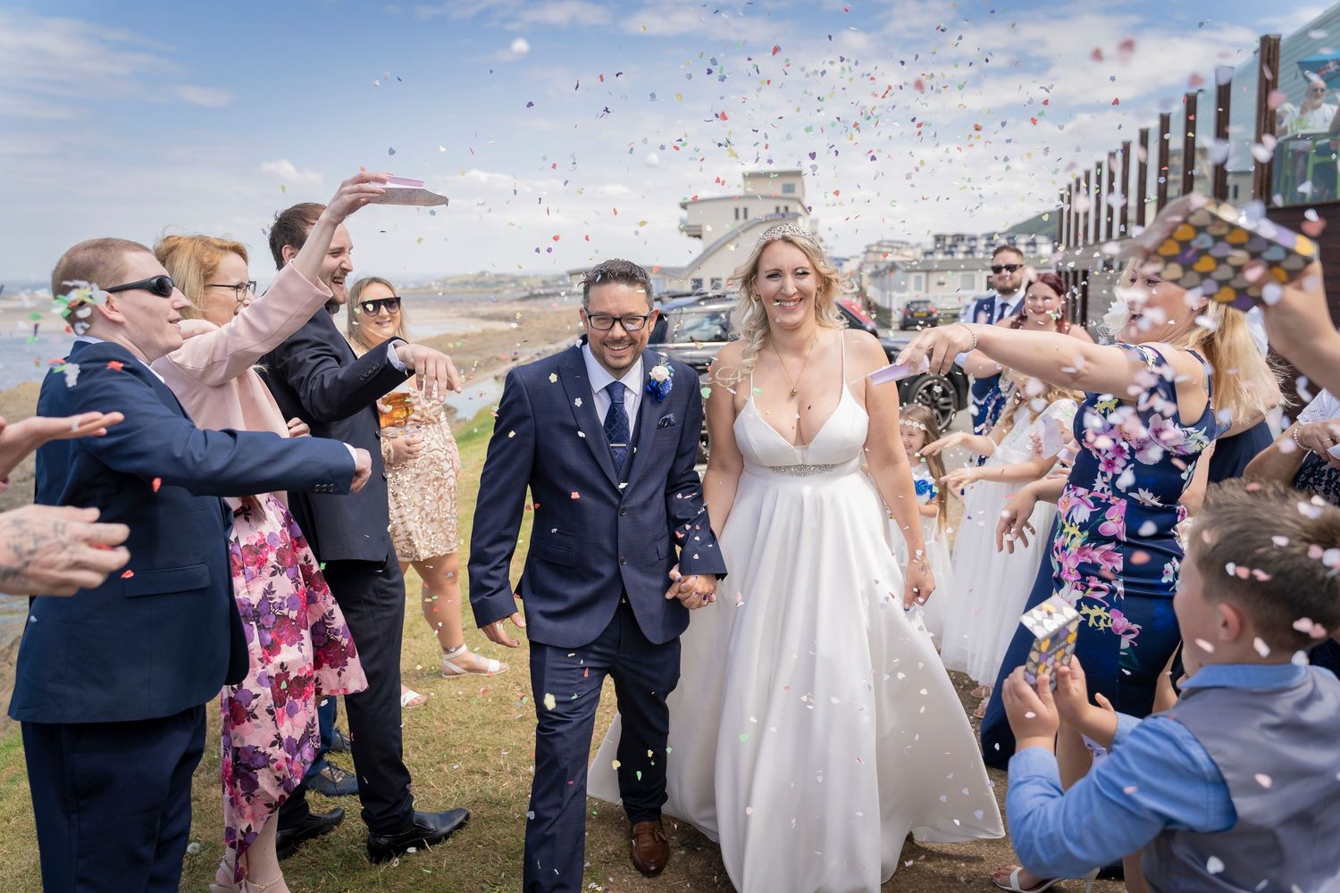 Happy newlyweds walk through shower of confetti with celebrating guests on sunny boardwalk.
