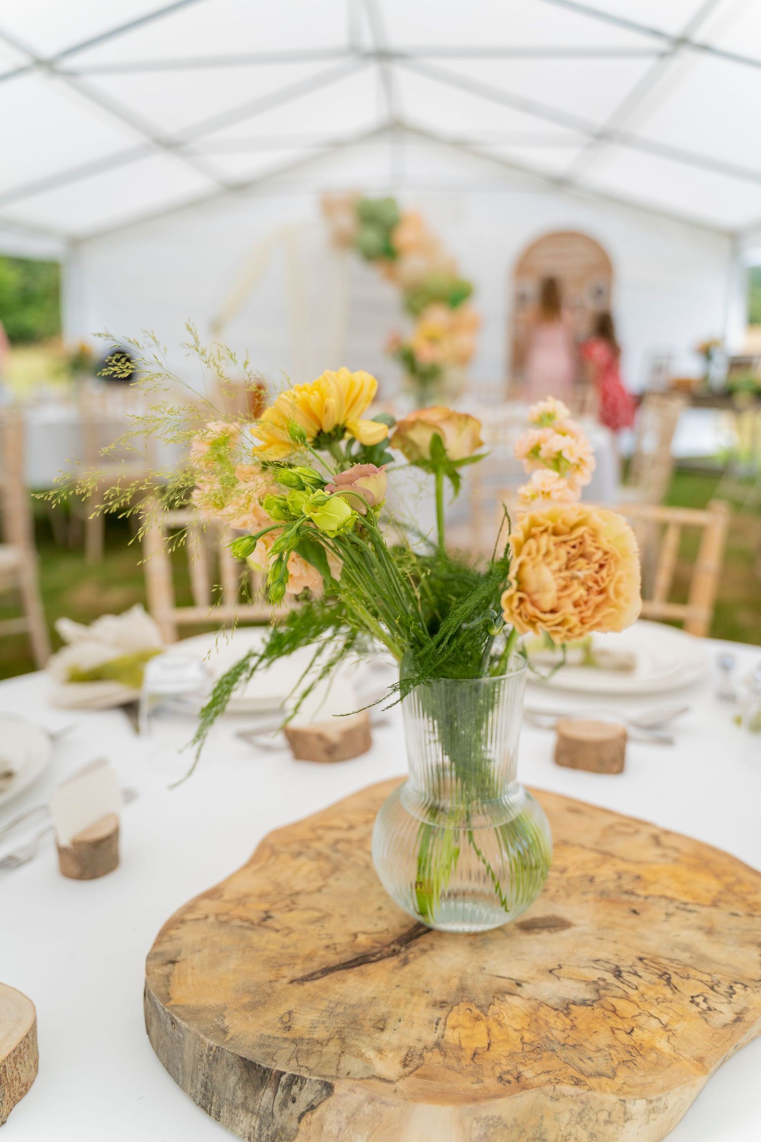 Close-up of yellow floral centerpiece in glass vase on natural wood slice at outdoor wedding reception.