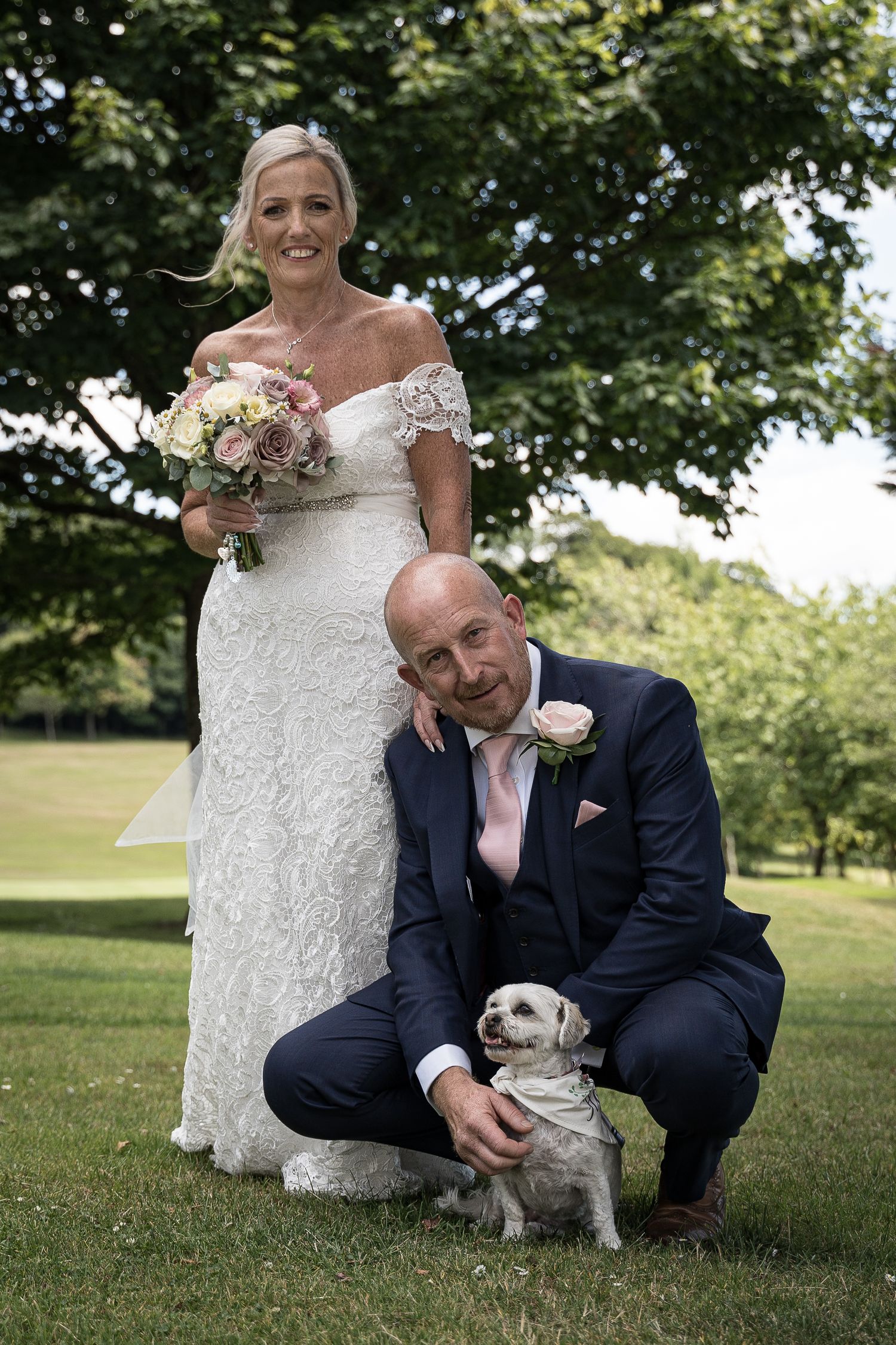 A wedding couple poses outdoors with their small pug dog on a grassy lawn. Elfordleigh Hotel Wedding. Devon Wedding Photography