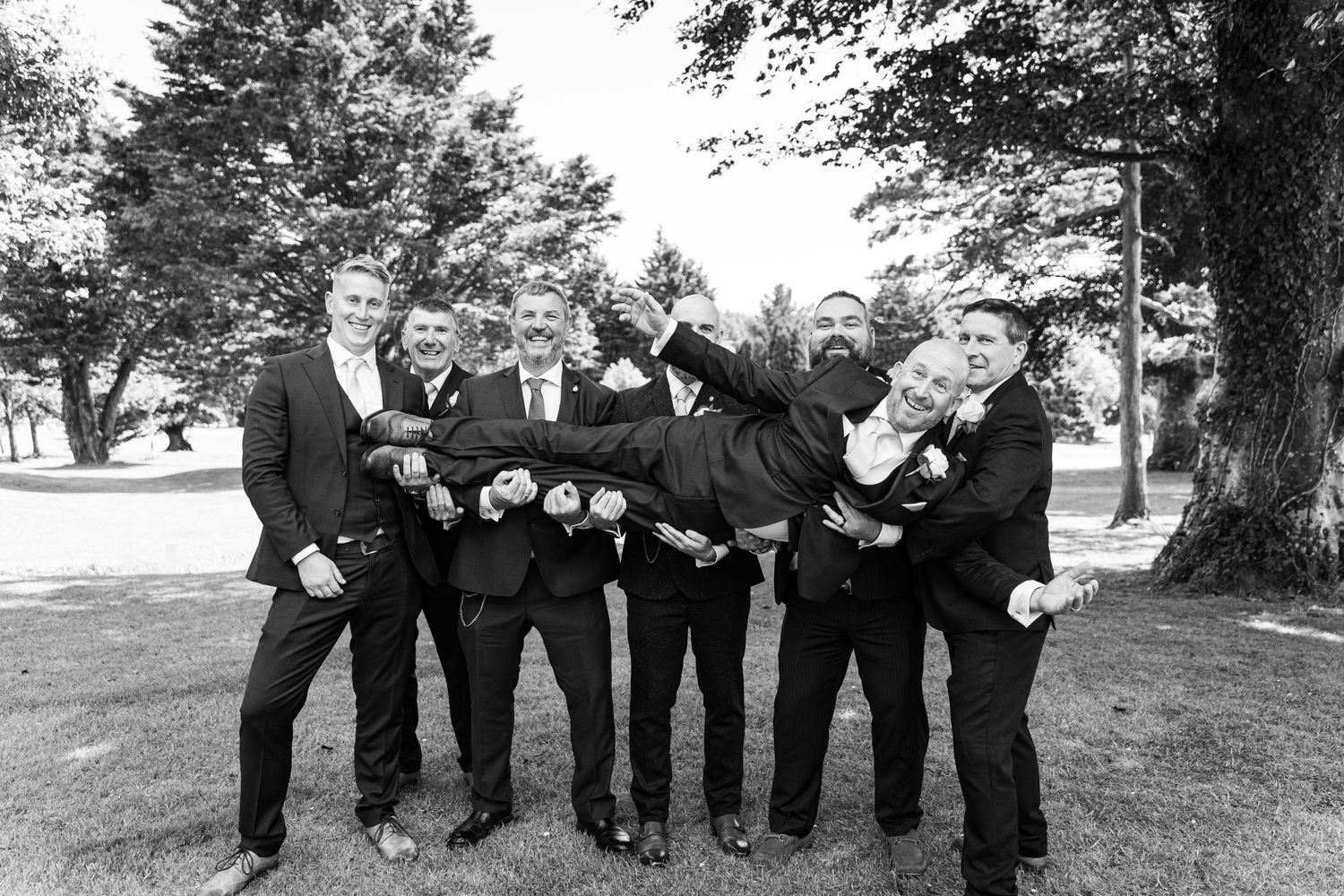 Groomsmen strike playful poses for black and white photo under trees.