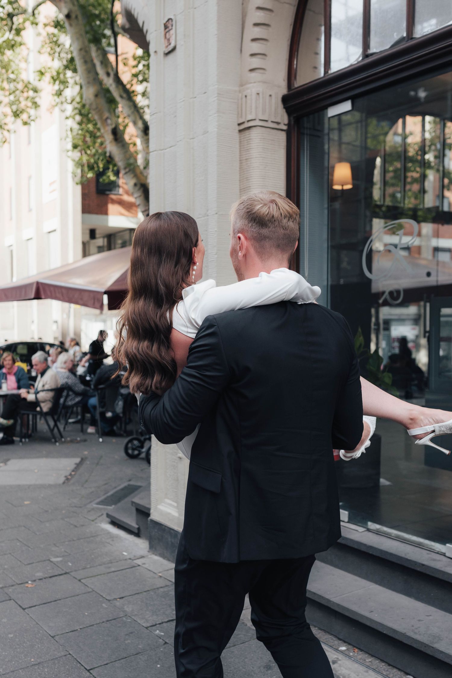 A couple embraces outside a city cafe, man in black suit with arm around woman with long dark wavy hair.