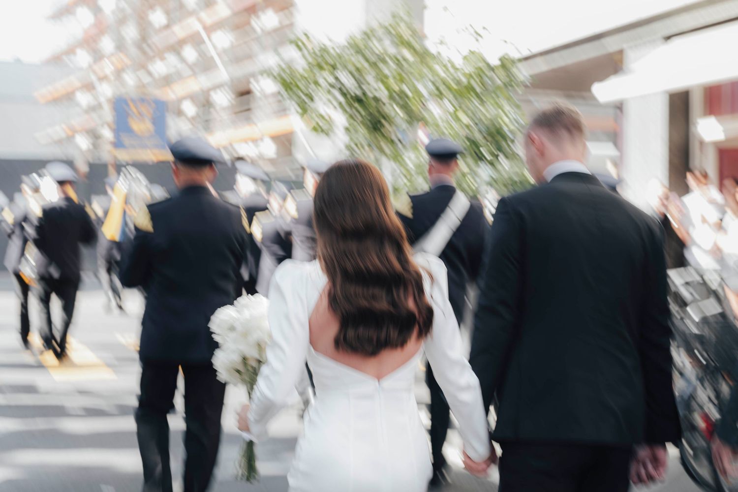 Bride and groom walking hand in hand outdoors, bride holding white bouquet, with uniformed officers in the background.