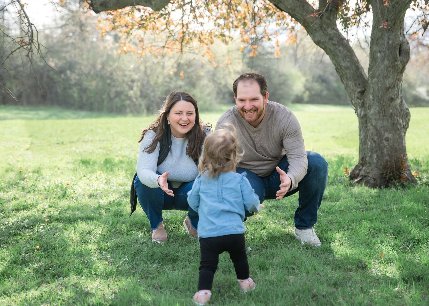 A joyful family with a toddler walking toward smiling parents outdoors under a tree on green grass.