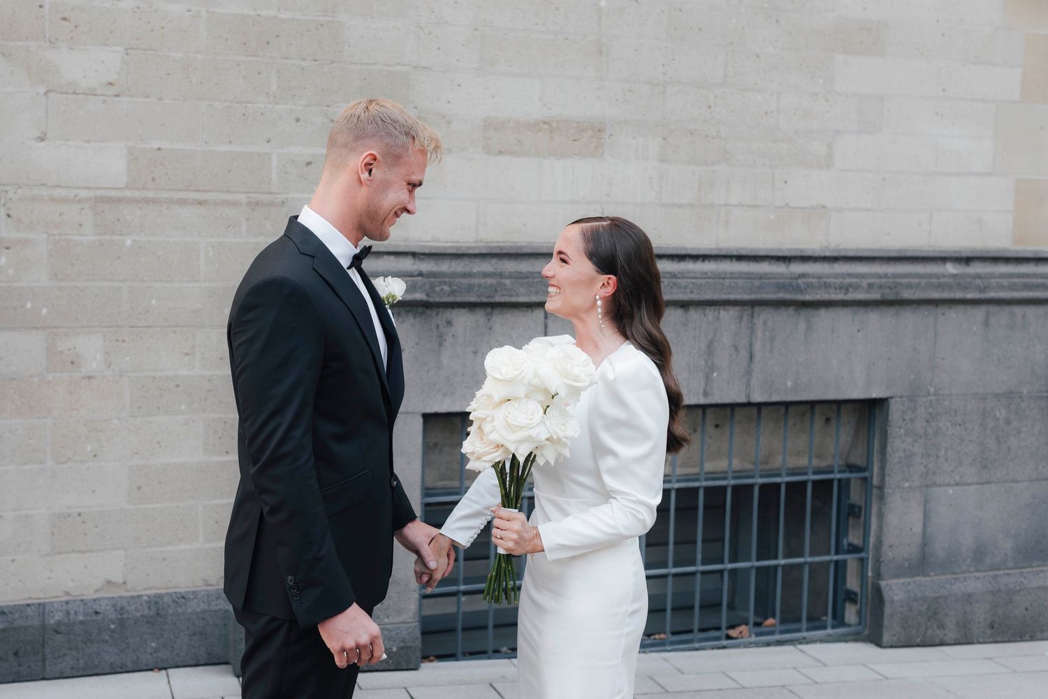 A bride and groom hold hands smiling at each other, she holds white peonies, both dressed in elegant wedding attire outdoors.