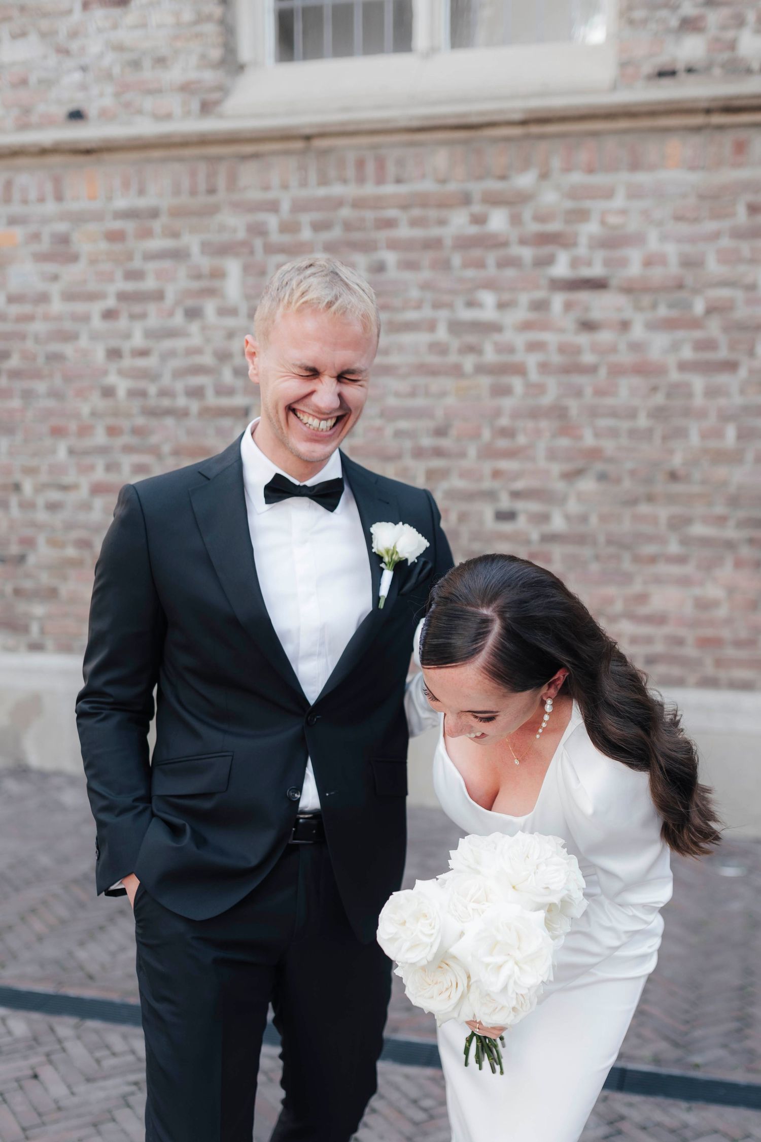 Bride in white gown holding white peony bouquet laughs with groom in black tuxedo with bow tie outdoors.