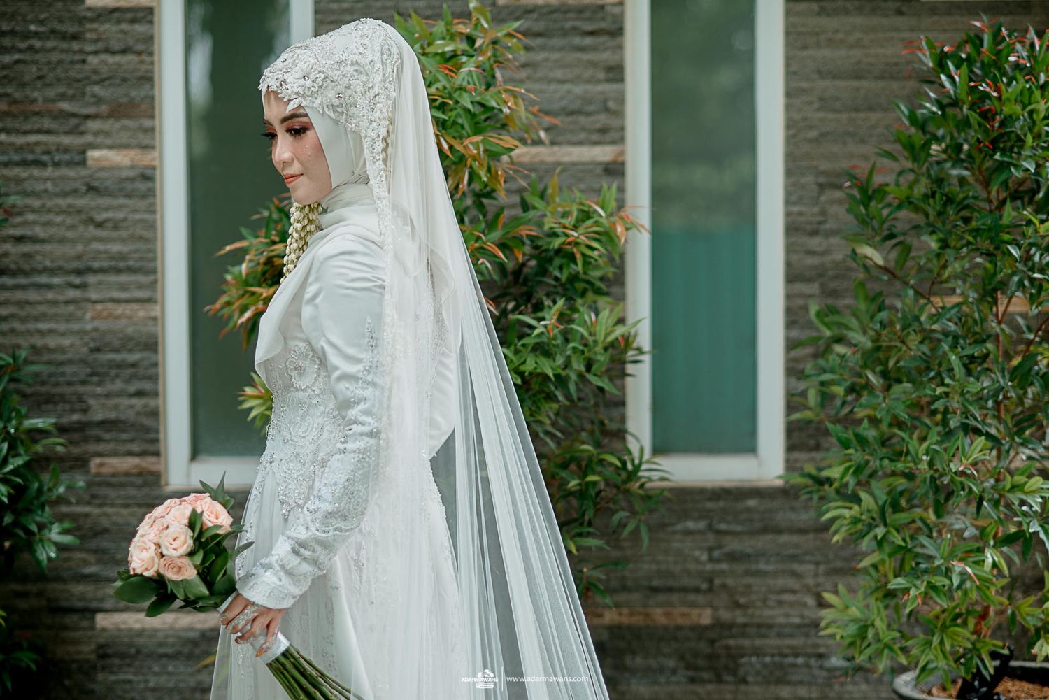 A bride in white Islamic wedding dress holds a bouquet against a modern building facade.