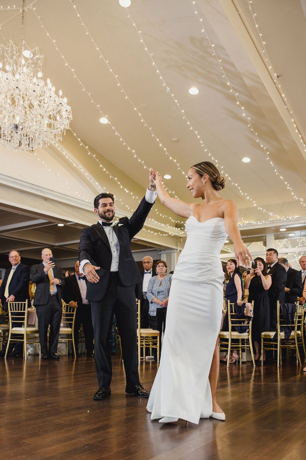 Bride and groom share a joyful first dance moment under sparkling string lights and chandeliers.