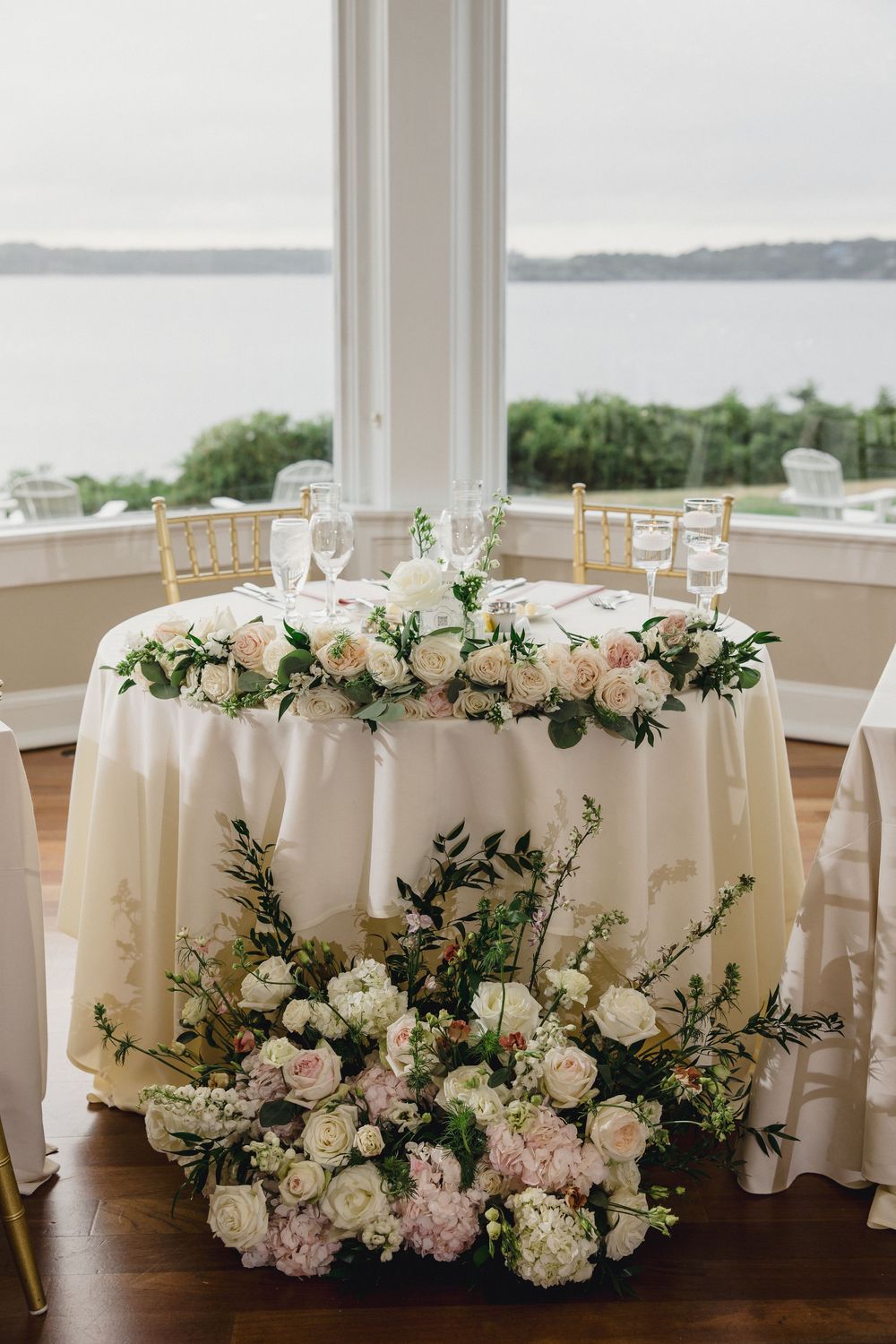 Elegant sweetheart table adorned with lush white and pink floral arrangements with waterfront backdrop. OceanCliff Hotel in Newport, RI