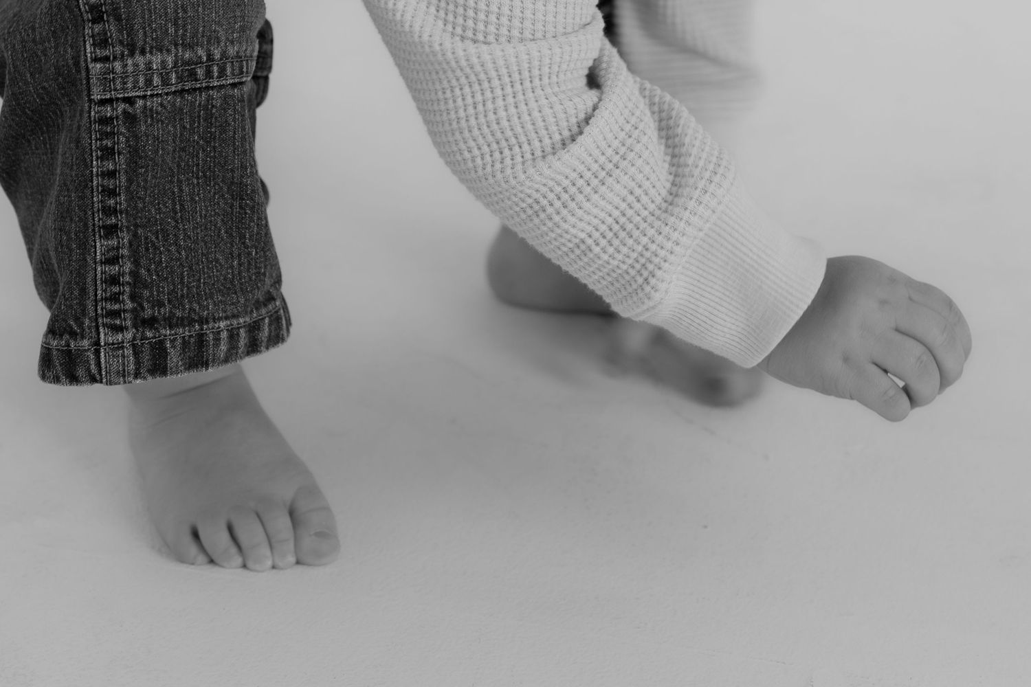 Black and white photo of two young children's bare feet standing on a white surface, wearing jeans and white pants.