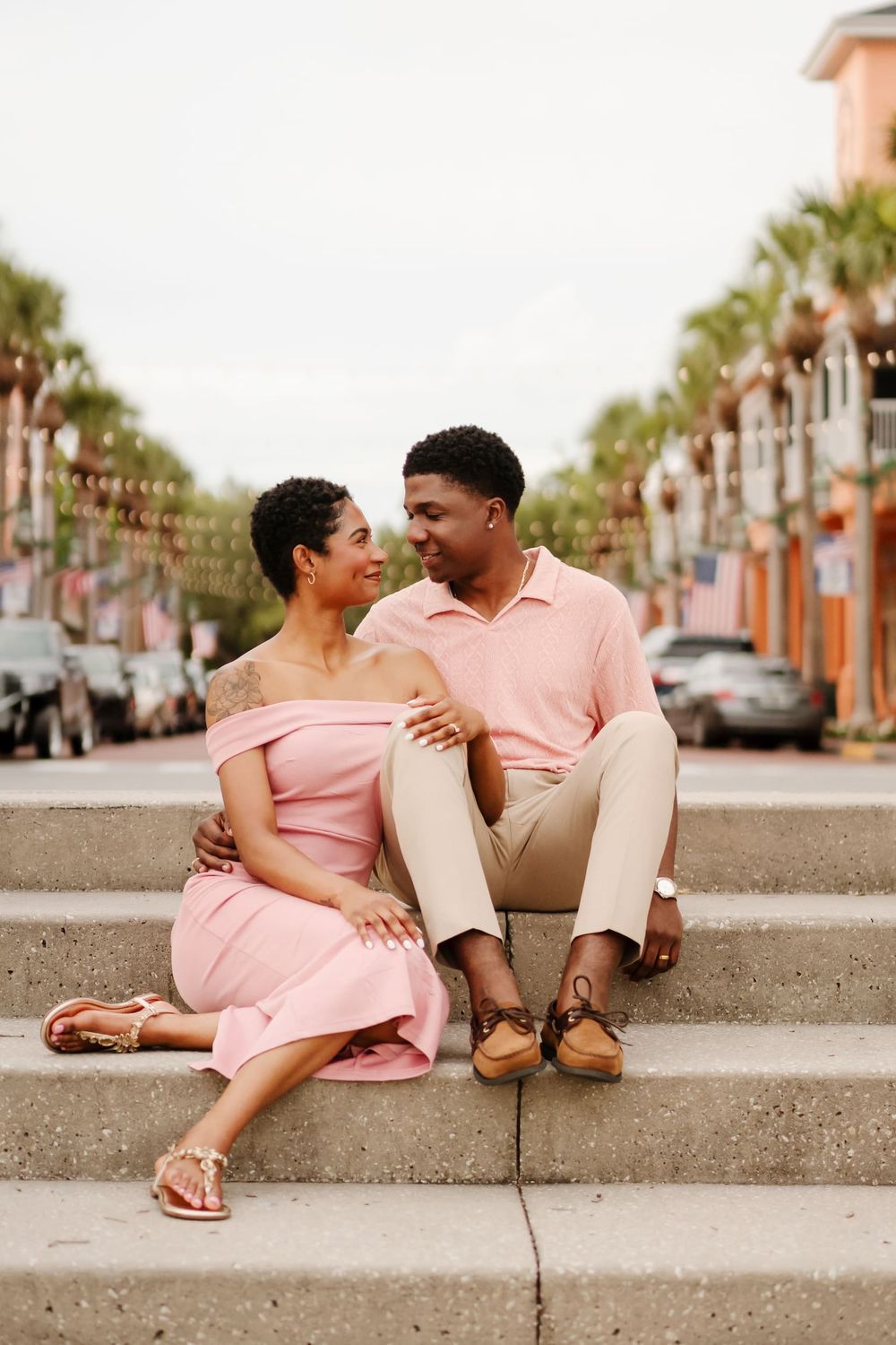 A couple in coordinating pink outfits sitting on steps, gazing at each other on a palm-lined street.
