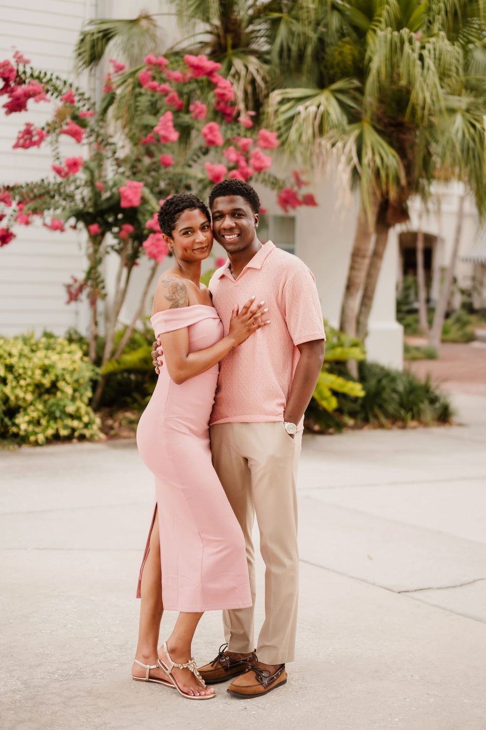 A couple in matching pink outfits pose together outdoors with tropical flowers and palm trees in the background.