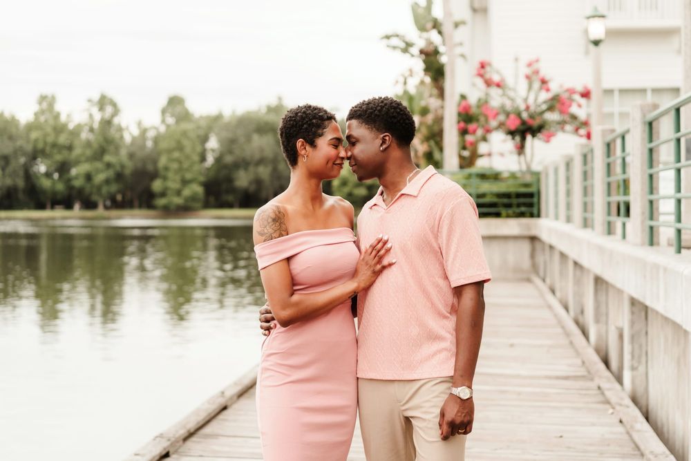 Couple in matching pink outfits sharing a romantic moment on a lakeside boardwalk surrounded by lush greenery.