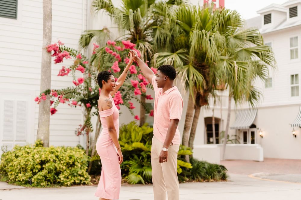 Couple dancing outdoors near tropical flowers and palm trees, woman in pink dress, man in pink shirt.