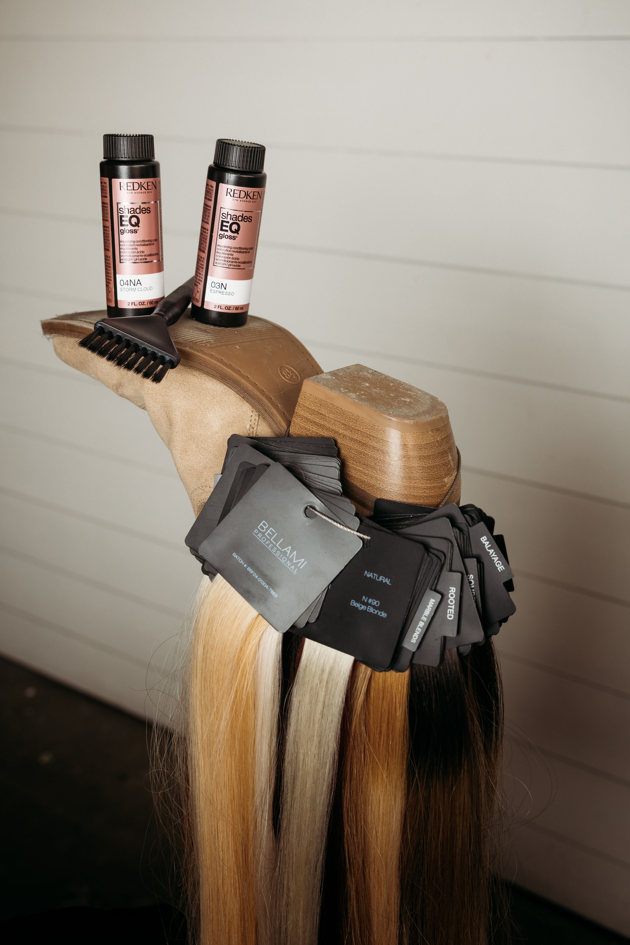 Redken EQ hair color bottles and Bellami hair extension swatches displayed on a wooden mannequin head stand.