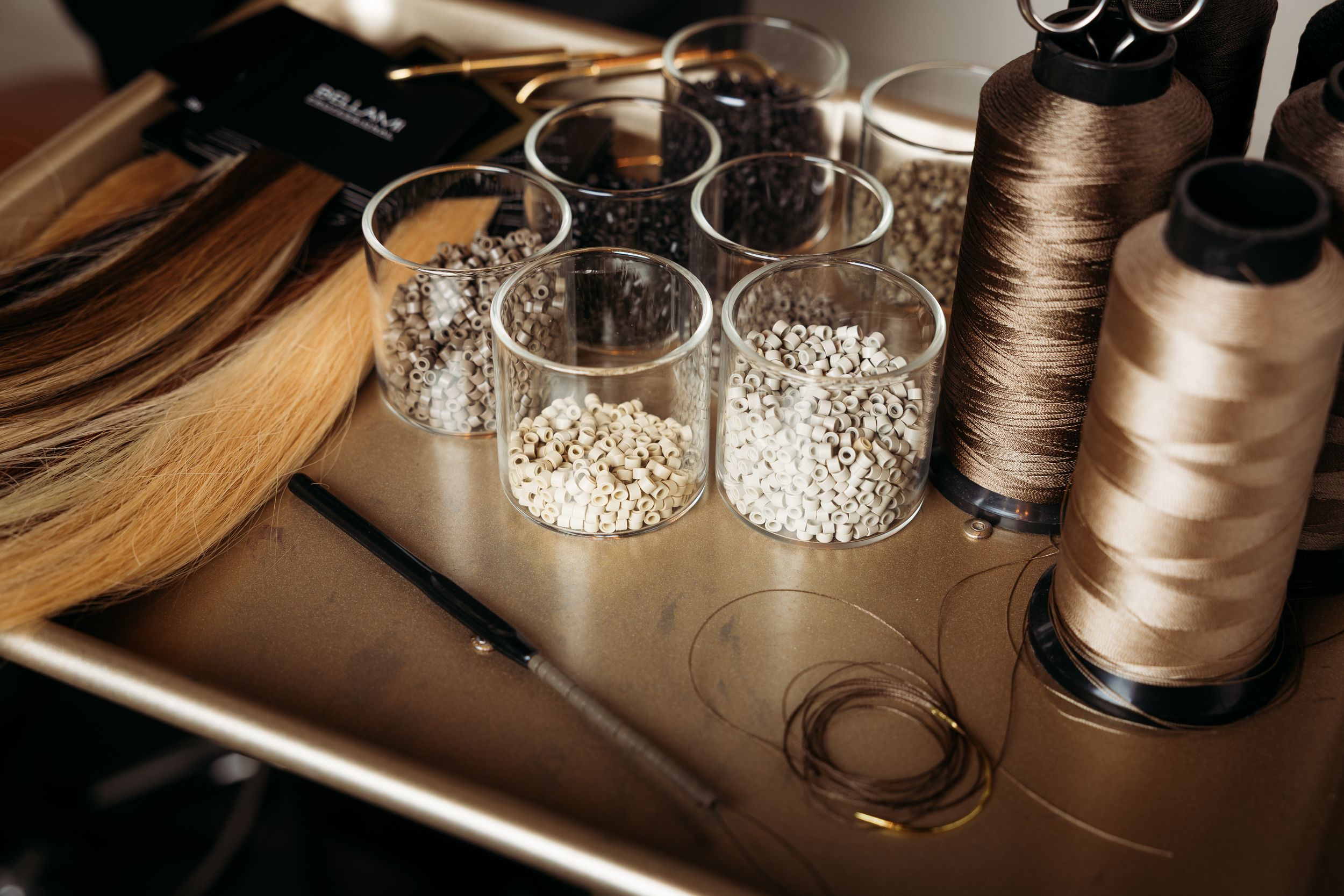 Hair extension tools with beads in glass jars, thread spools, and a needle on a golden tray.