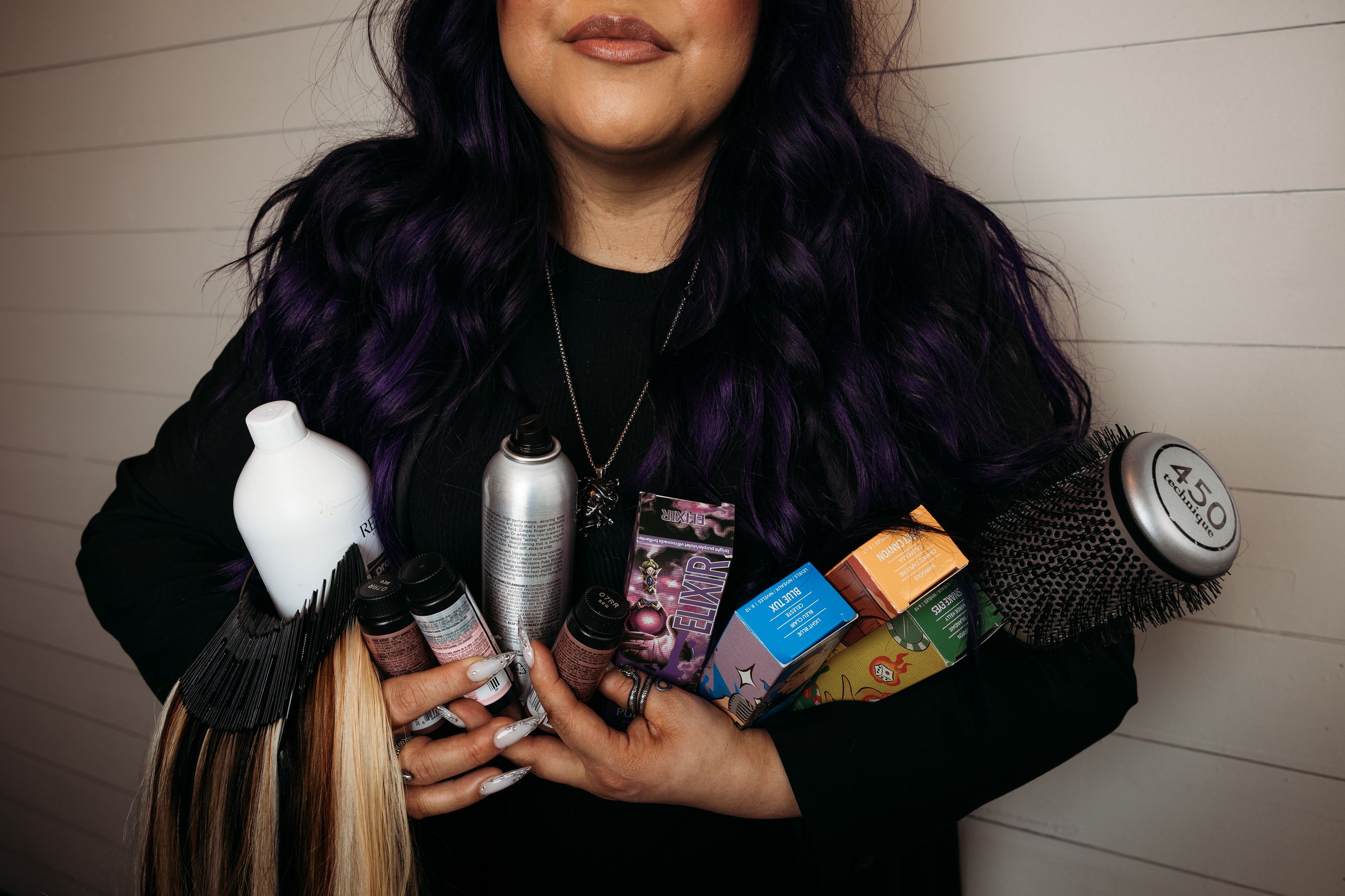 A hair stylist with purple hair holds styling tools, products, and hair extensions in a professional salon setting.