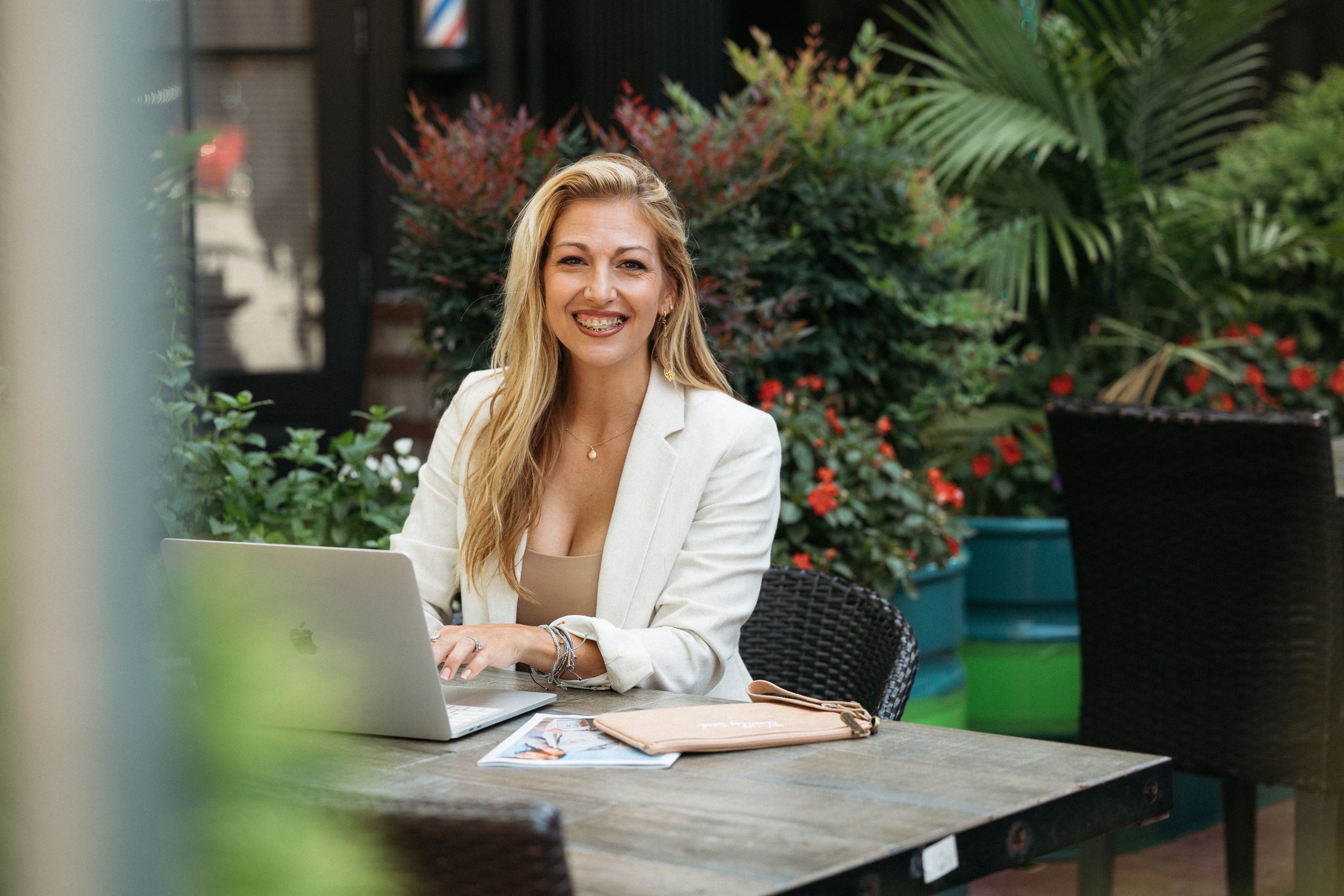 Professional woman in white blazer smiling while working on laptop at outdoor cafe surrounded by greenery.