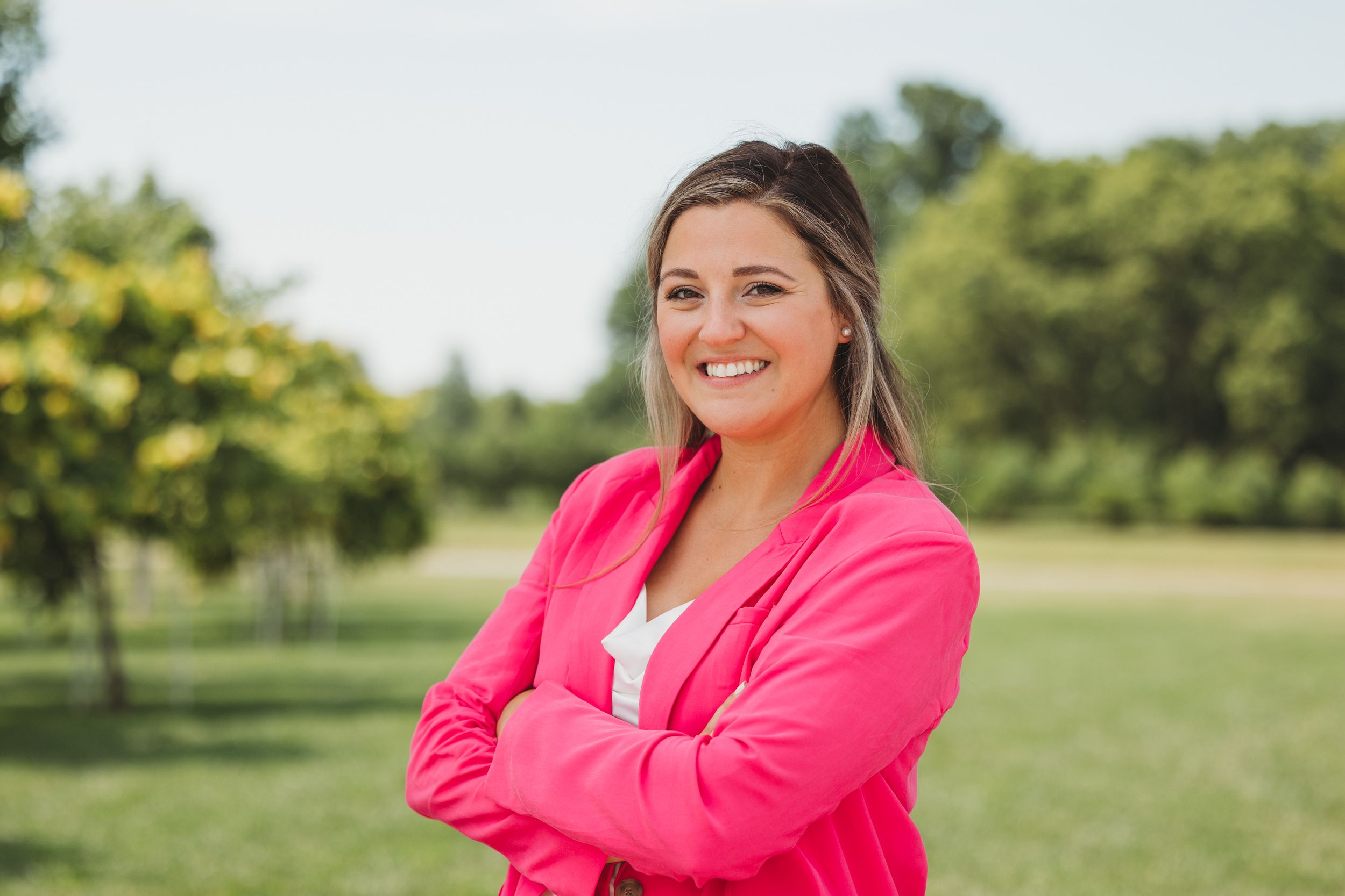 Smiling woman in pink blazer with arms crossed standing outdoors in a lush green park setting.