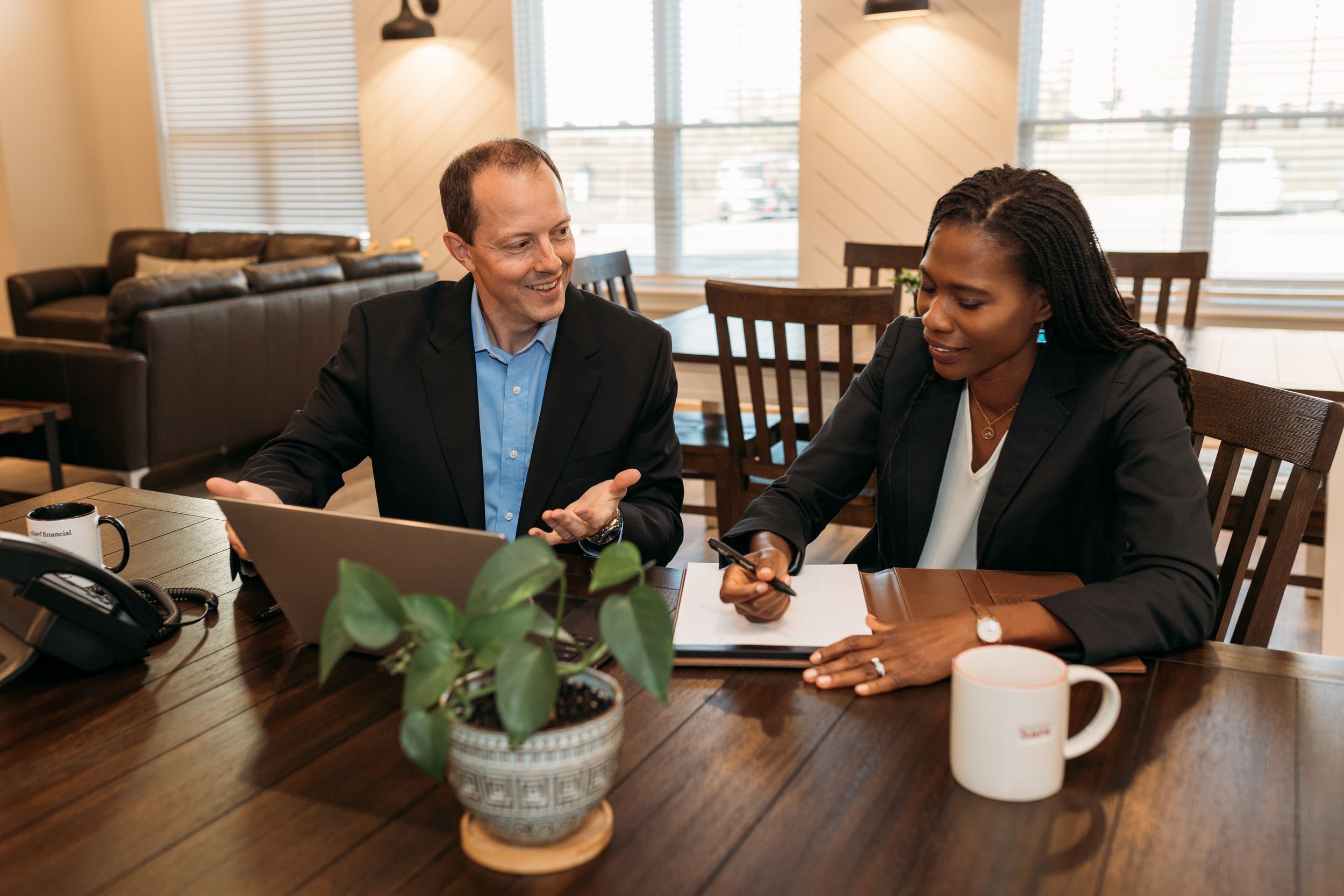 Two business professionals collaborating over a laptop and documents at a wooden table in a modern office setting.