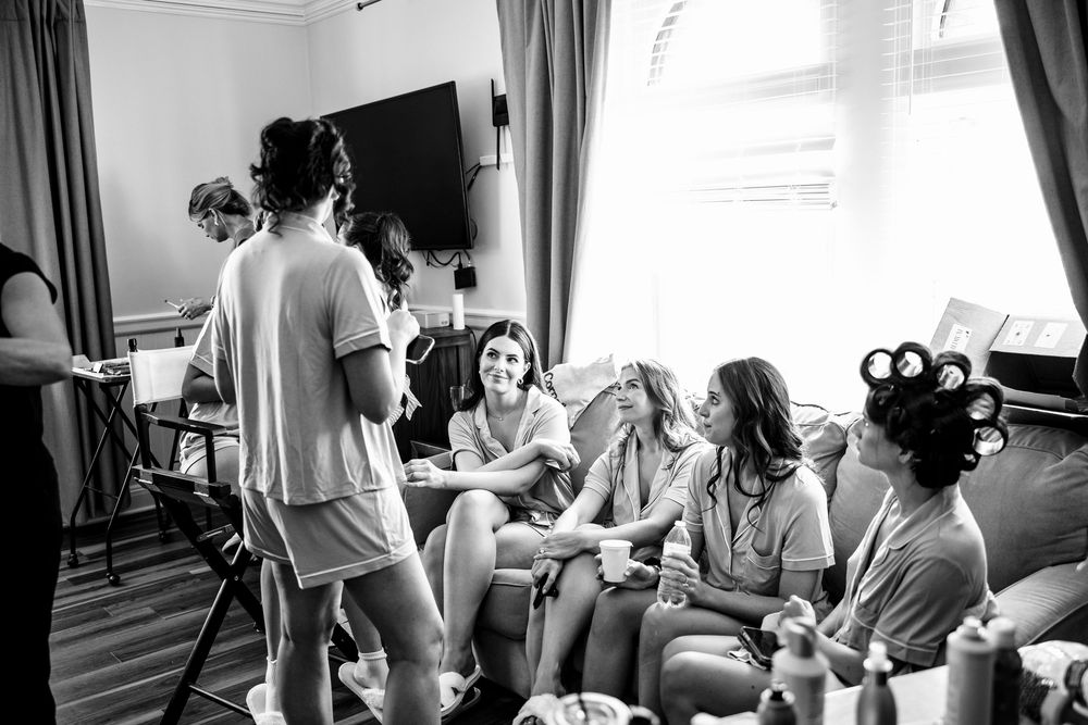 Black and white photo of bridesmaids getting ready, laughing together with hair rollers in a bright room.