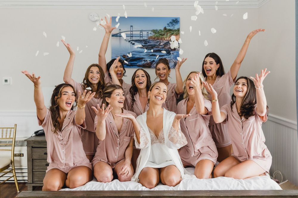 Bride and bridesmaids in mauve robes celebrate on a bed tossing white flower petals in the air. OceanCliff Newport RI