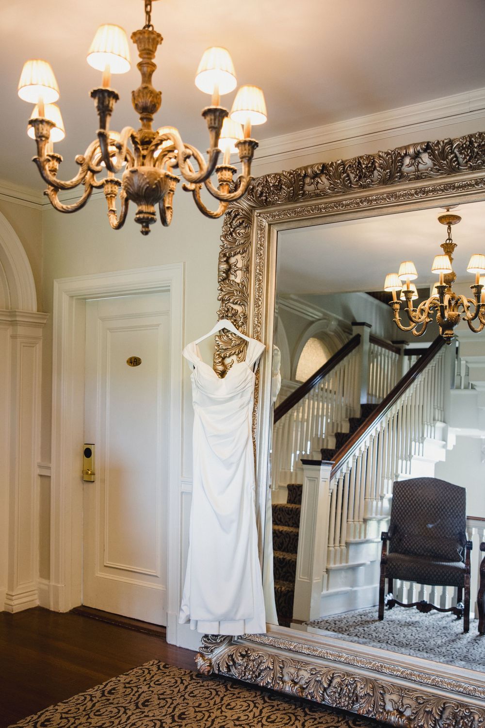 Elegant wedding dress hanging in a grand foyer with ornate gold chandelier and large decorative mirror. Ocean cliff RI