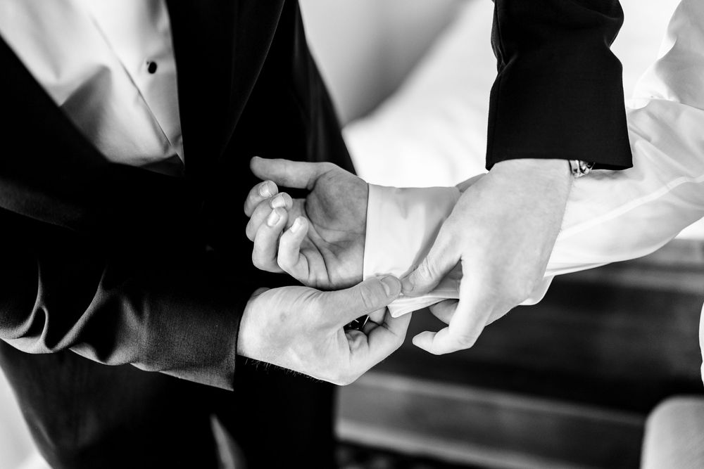 Black and white photo of groomsmen fastening cufflinks on a tuxedo shirt before a wedding ceremony.