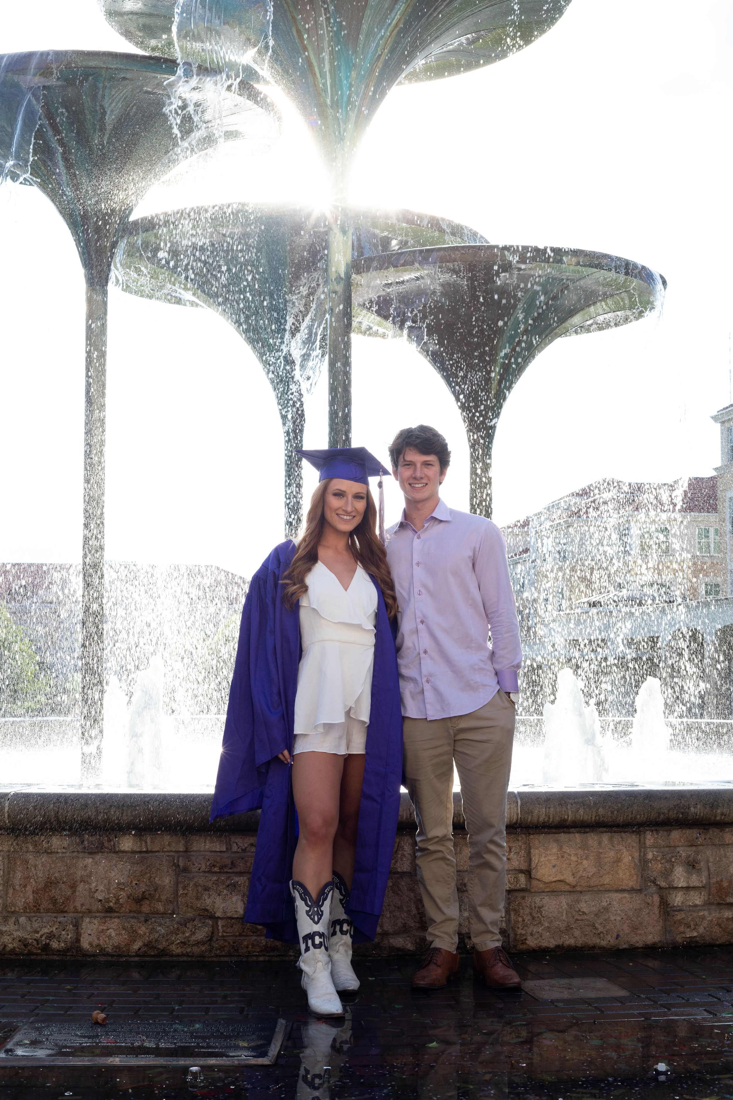 TCU graduate in purple cap and gown posing with partner in front of sunlit campus fountain after graduation ceremony.