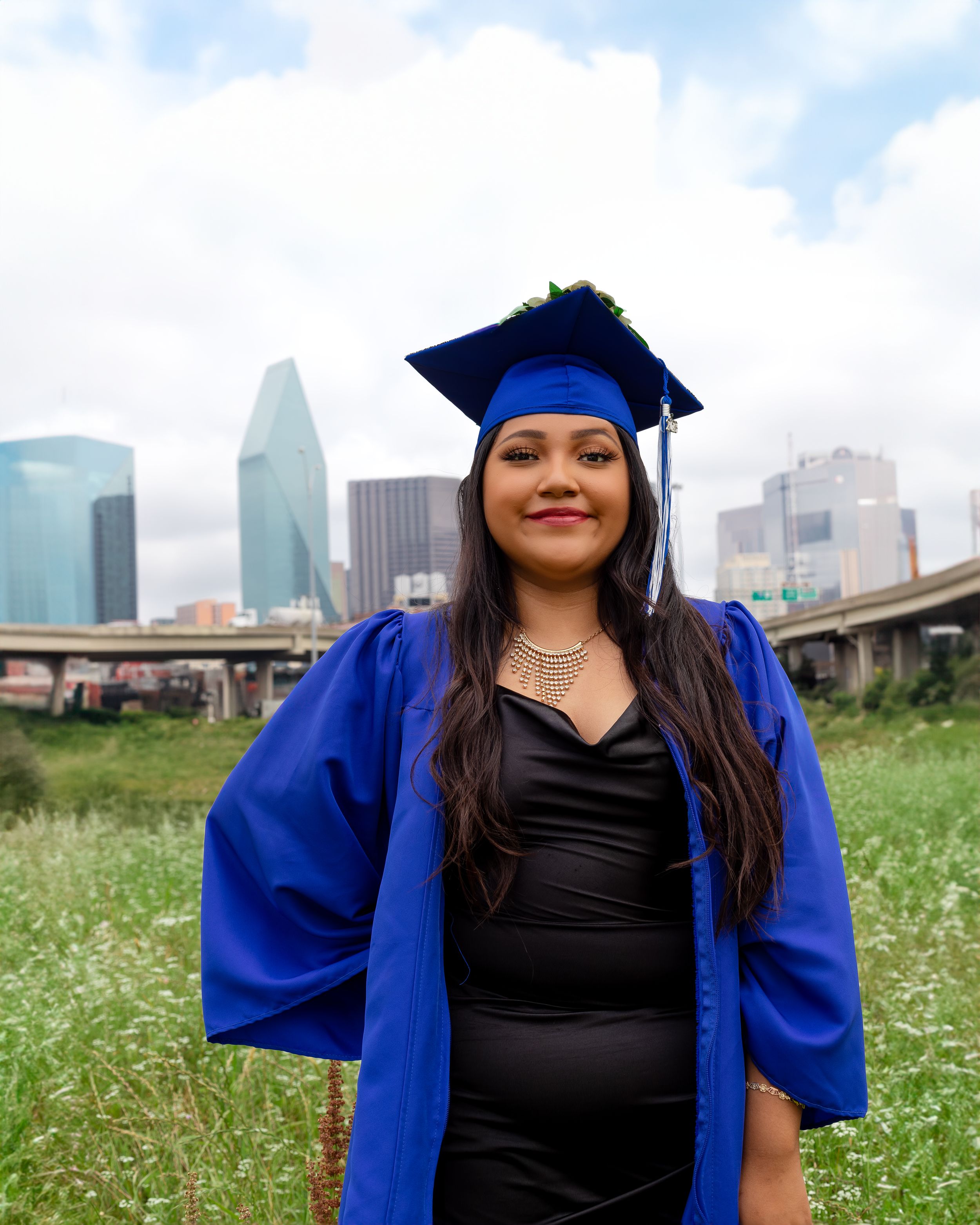 College graduate in blue cap and gown posing outdoors with Dallas skyline backdrop, celebrating academic achievement.