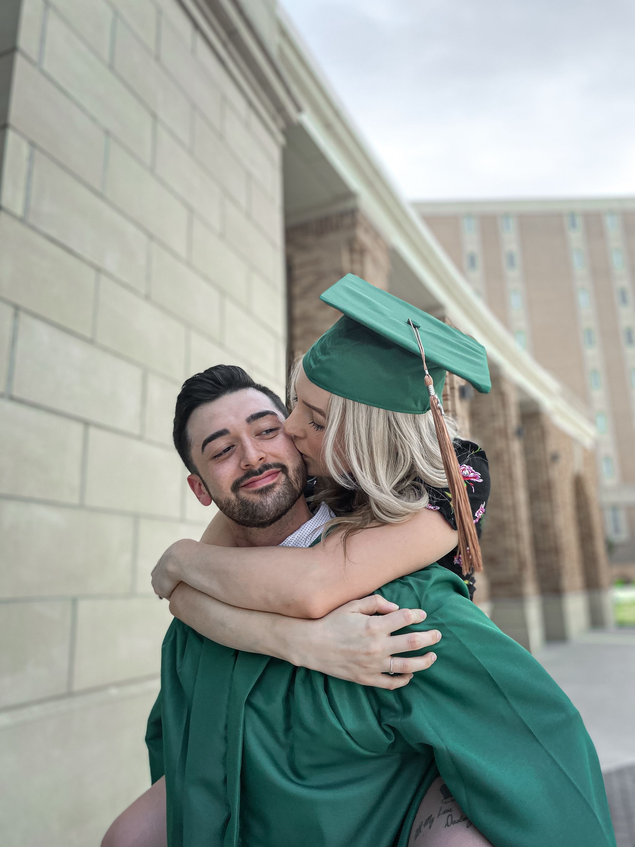 Graduate in green cap and gown sharing a celebratory embrace and kiss outside campus after graduation ceremony.