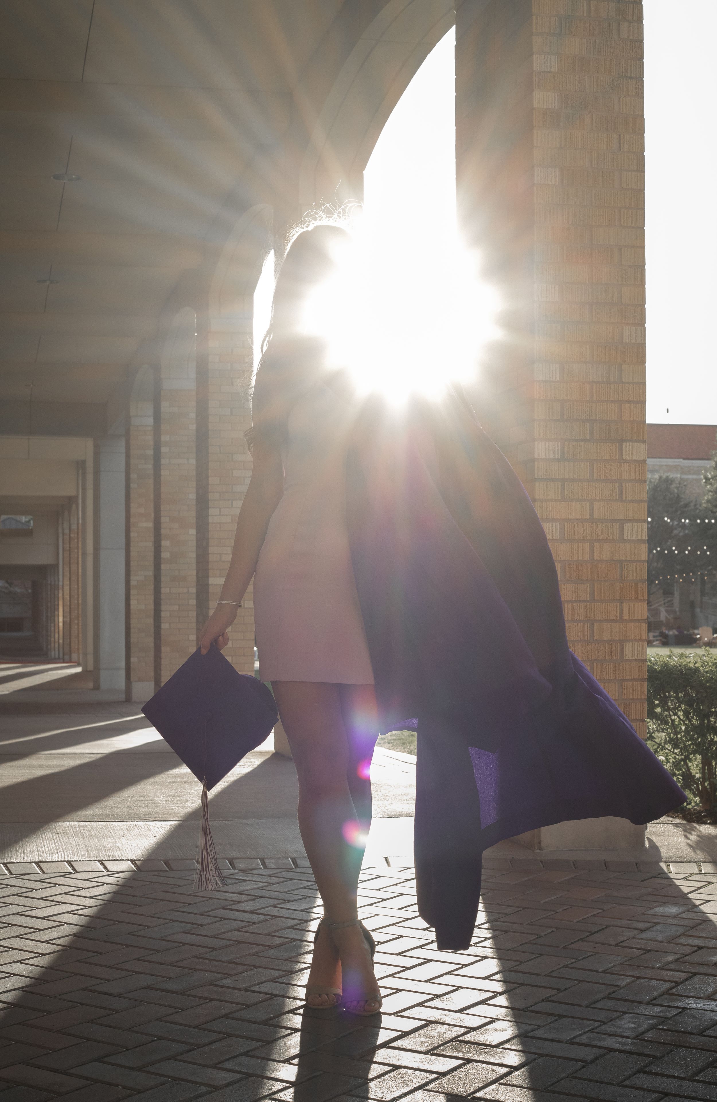 Graduate holding cap and gown in sunlit campus walkway with dramatic lens flare, celebrating graduation achievement.