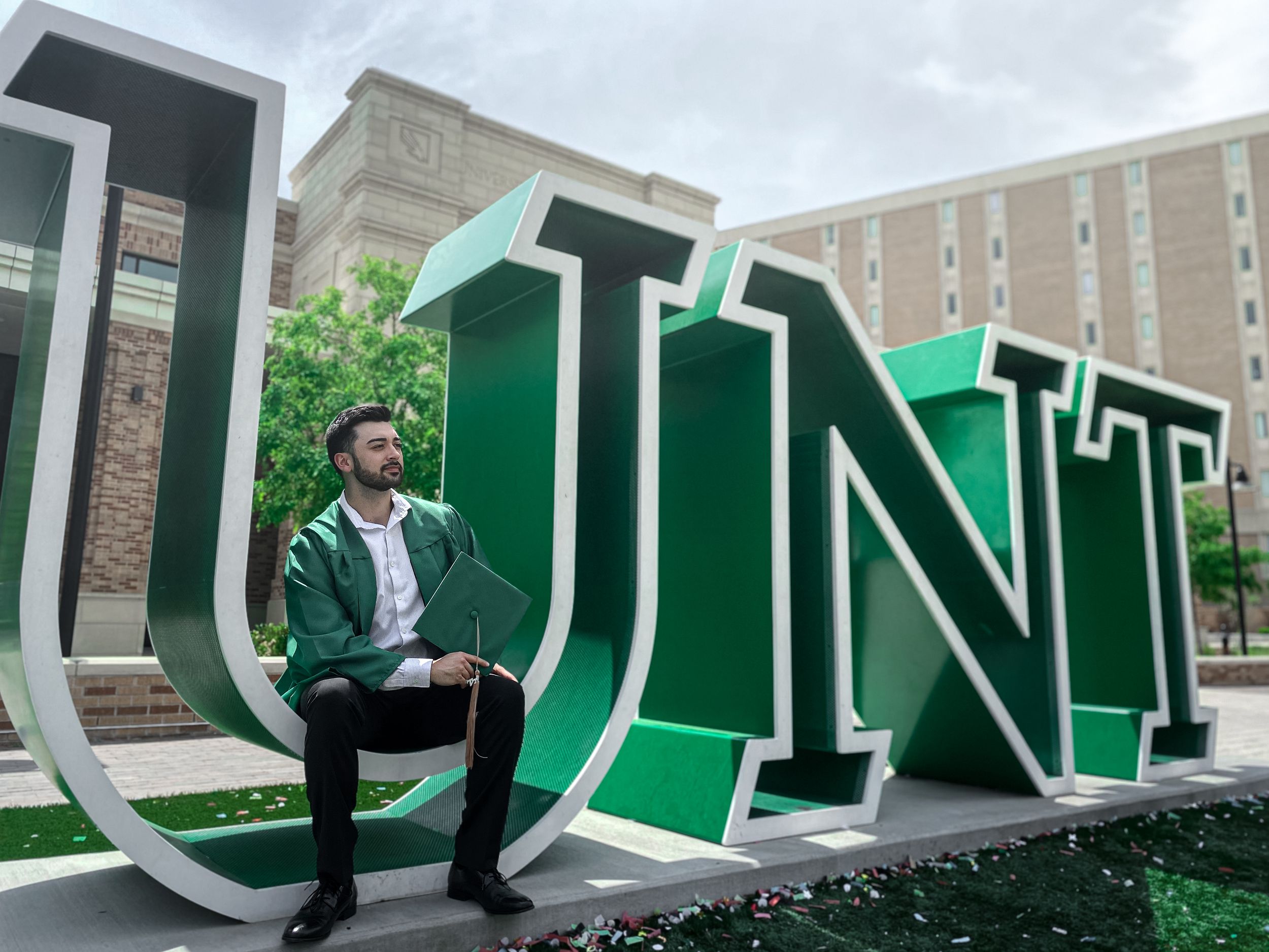 University of North Texas graduate posing beside the iconic green UNT sign on campus, celebrating graduation on a sunny day.