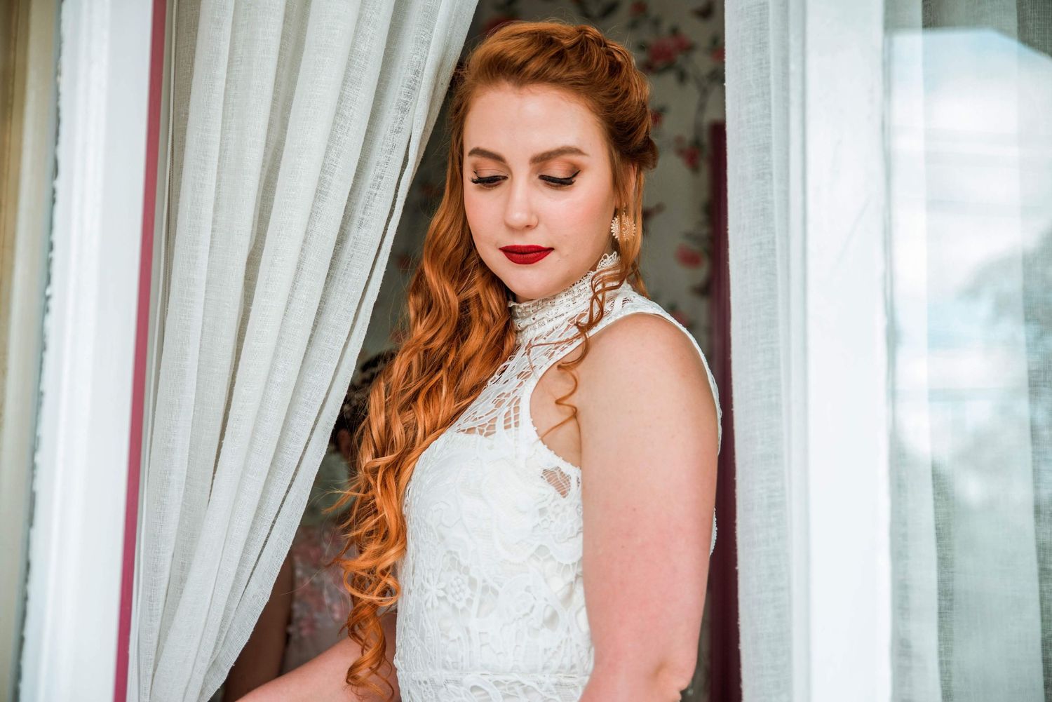 A portrait of someone in a white lace wedding dress standing by sheer curtains.