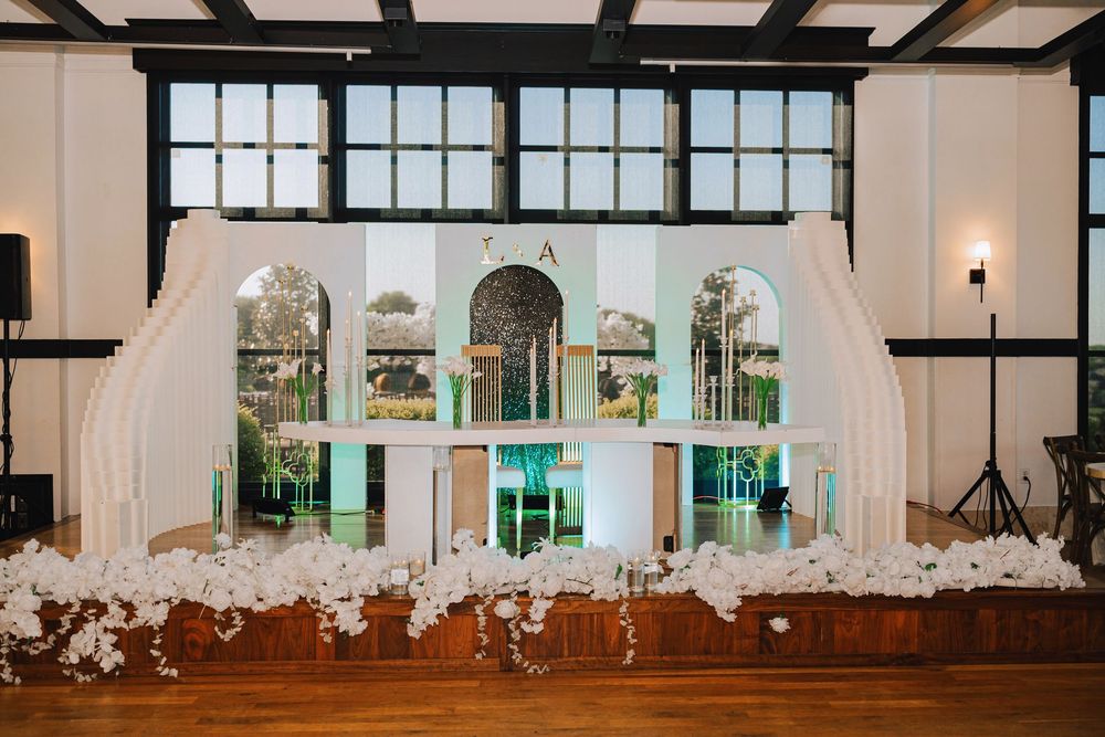 Elegant wedding reception venue with white arch backdrop, floral arrangements, and large windows in a grand ballroom.