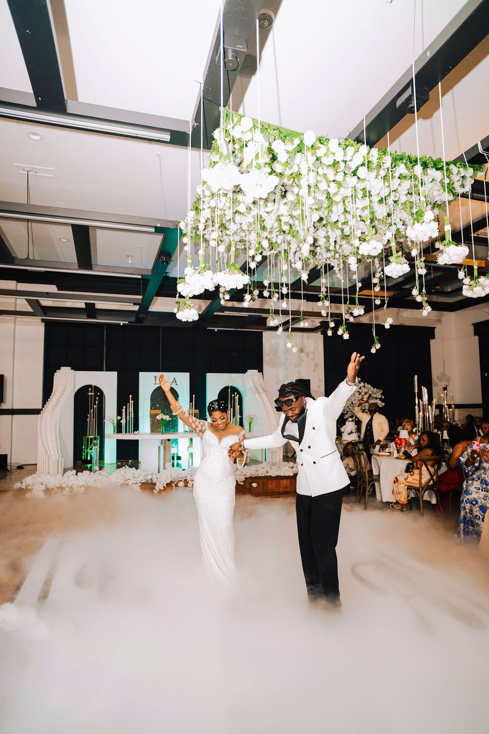 Bride and groom share first dance surrounded by fog under floral chandelier at elegant wedding reception.