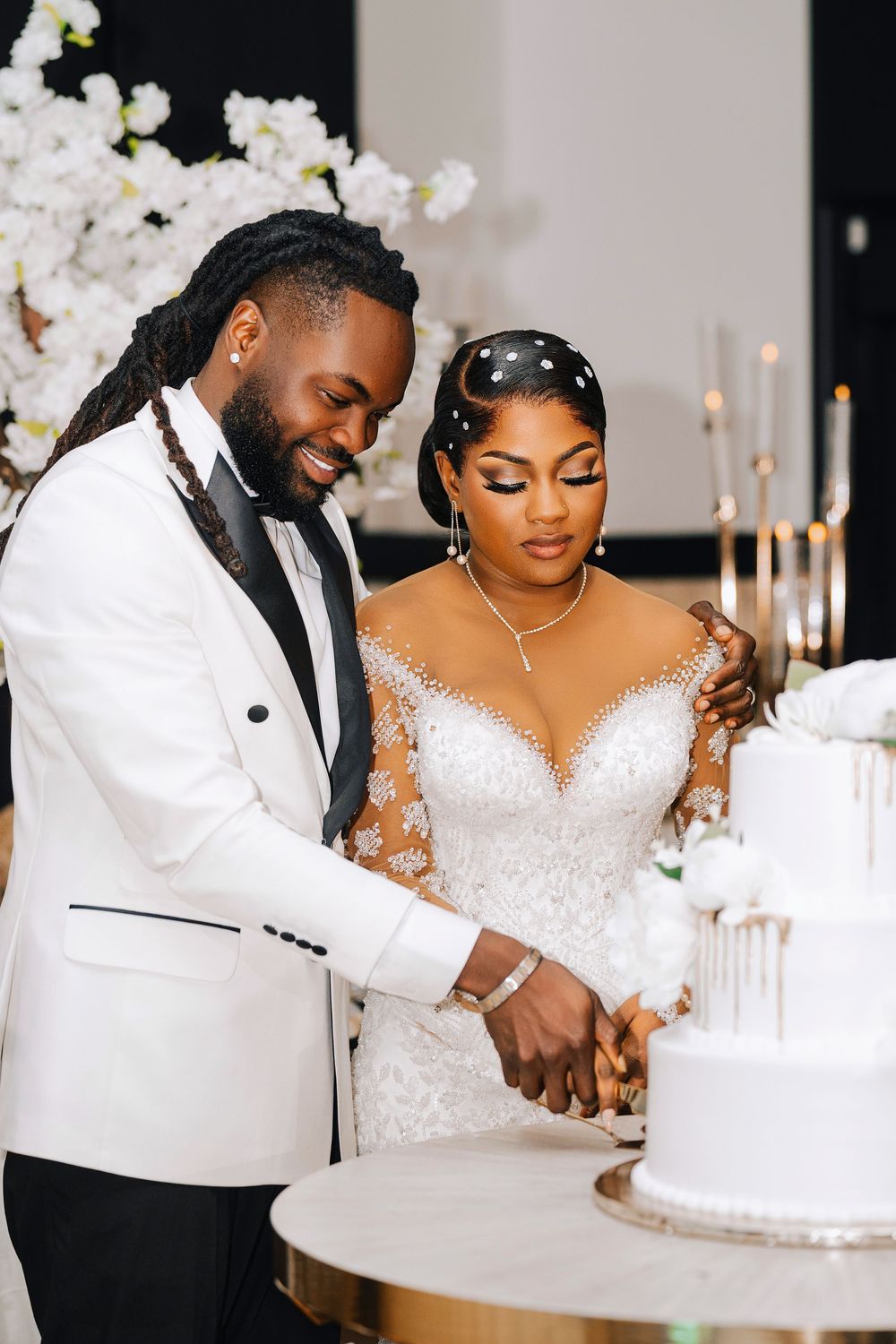 Bride and groom cutting white wedding cake, wearing elegant white attire with floral decor in background.