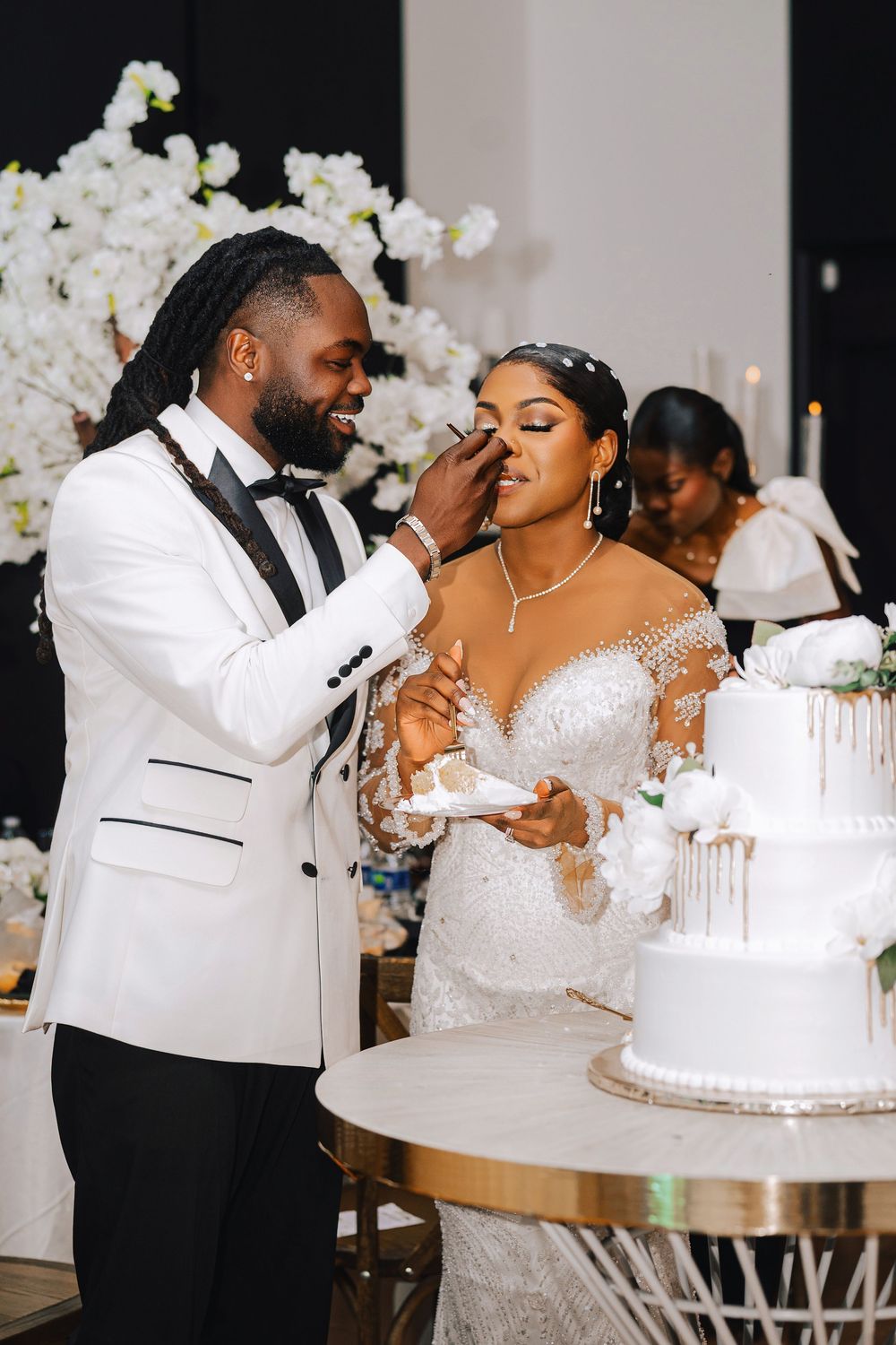 Groom in white tuxedo playfully smearing wedding cake on bride's face during elegant reception.