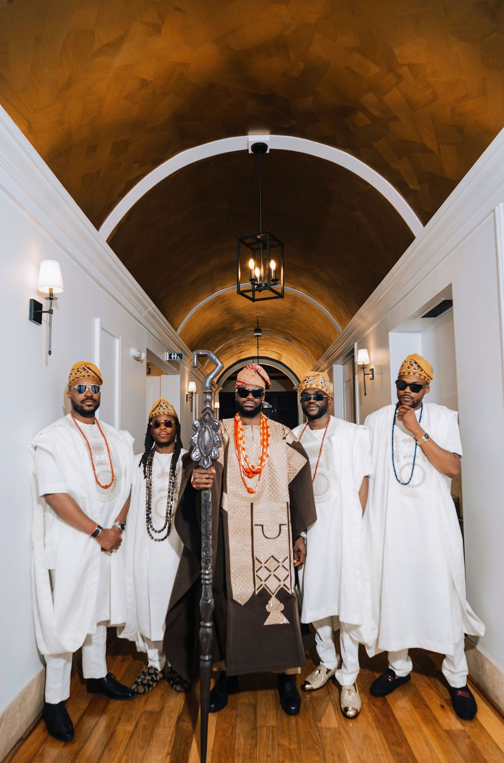 Five men in traditional West African white attire and colorful beaded accessories pose in an elegant arched hallway.