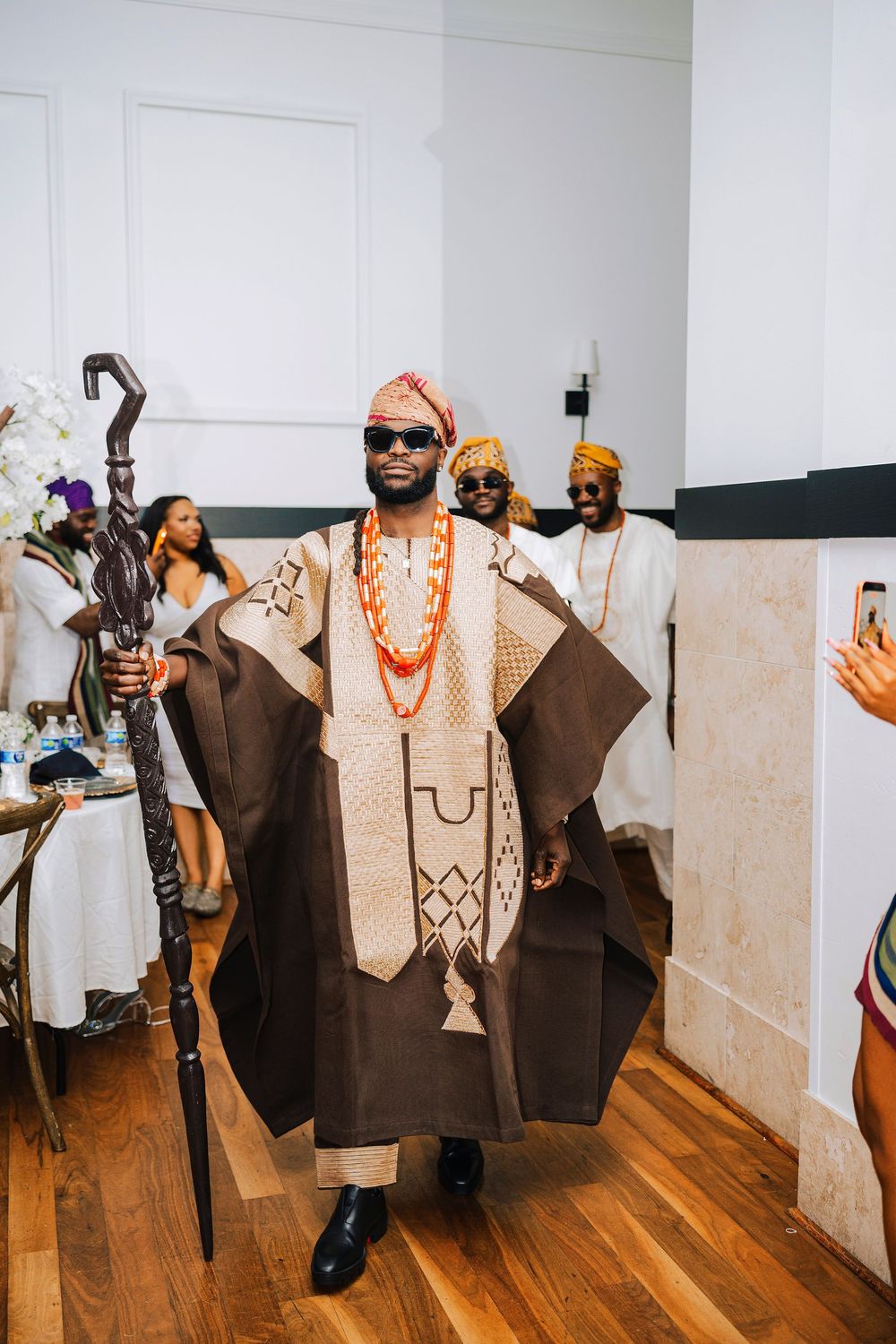 Man in traditional African agbada attire with coral beads and staff at a cultural ceremony.