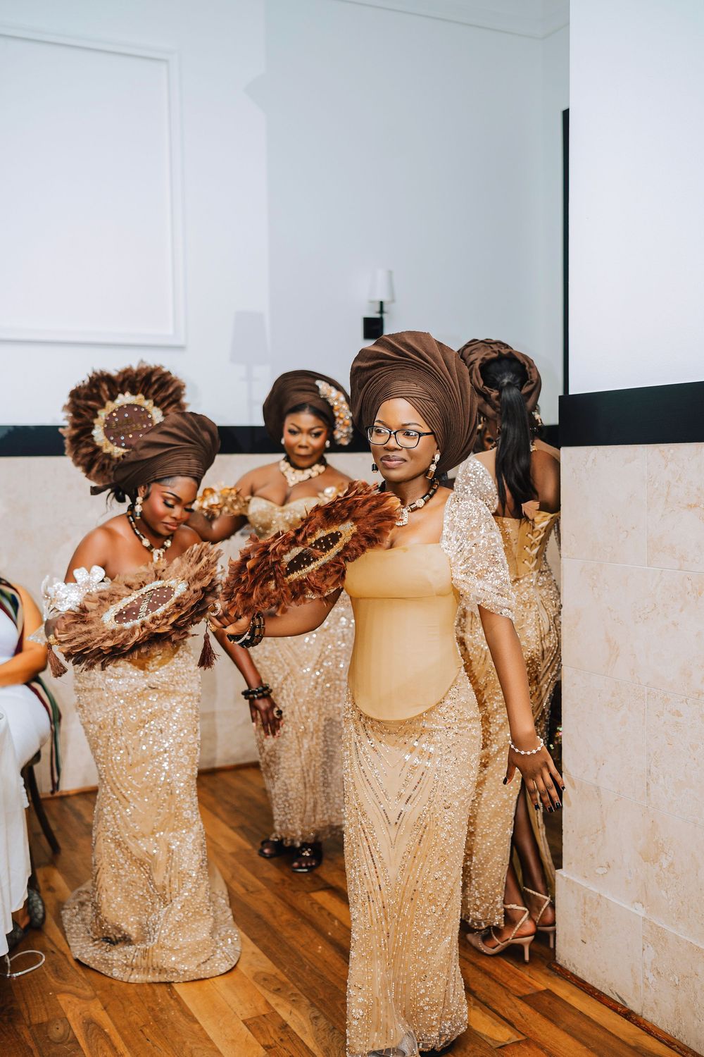 Bridesmaids in gold sequin dresses and brown head wraps hold feather fans at a traditional African wedding ceremony.