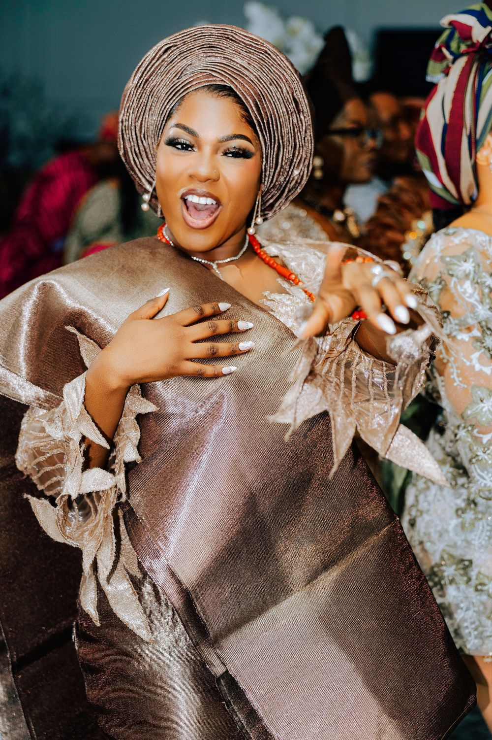 Woman in elegant gold Nigerian traditional attire with gele headwrap and coral beads celebrating at a wedding.