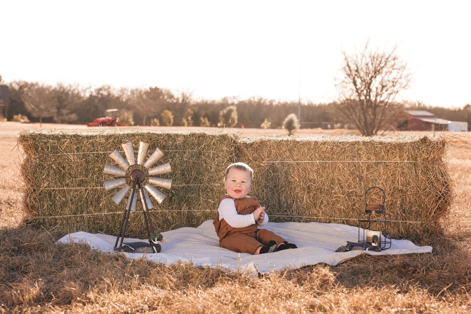 Baby sitting on a blanket in front of hay bales with a windmill decoration on a sunny farm.