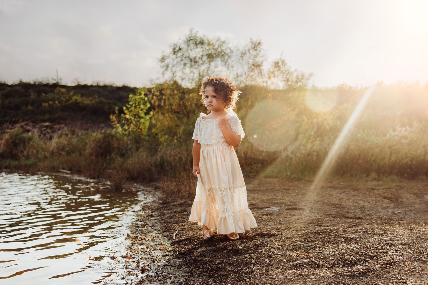 A young girl in a white dress stands by a sunlit riverbank, bathed in golden hour light.